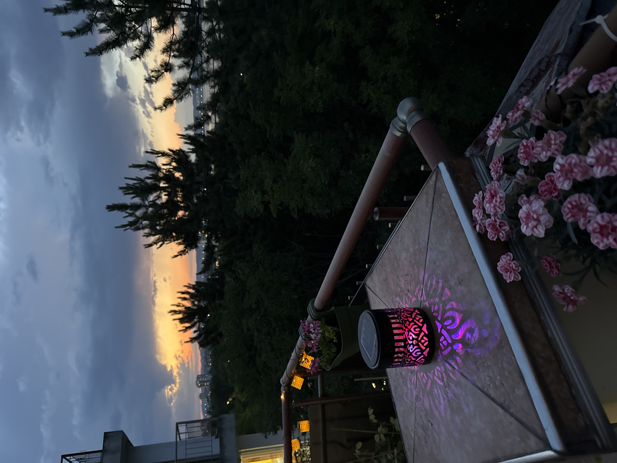 The image depicts a balcony overlooking a cityscape and a body of water, likely the bay of Burgas, Bulgaria, during twilight. The sky is a mix of dark clouds and hues of orange and yellow from the setting sun. Pine trees are prominent in the foreground, silhouetted against the sky. On the balcony, a table with a tiled surface holds a decorative solar lantern casting intricate purple patterns onto the table. Nearby, a pot contains pink flowers. A railing with wooden and metal elements runs along the edge of the balcony, and strings of small, square-shaped lights are visible. In the distance, city lights twinkle, and buildings are faintly visible. The overall atmosphere is serene and tranquil.