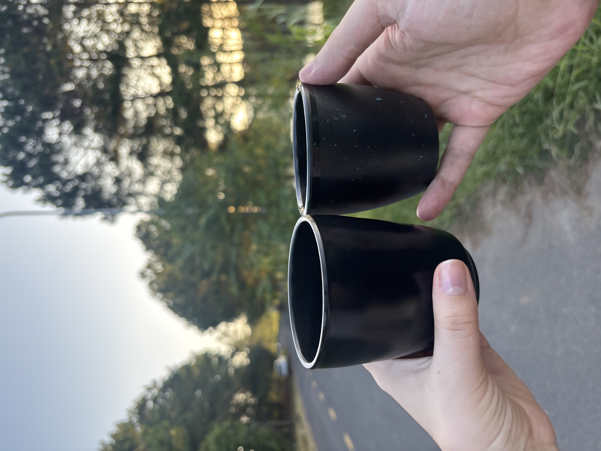 Two hands hold black cups in the foreground, with a blurred outdoor scene in the background. The left hand, with fair skin and short fingernails, holds a slightly larger, smooth black cup. The right hand, with tanned skin and visible fingerprints, holds a smaller, speckled black cup. Both cups are positioned as if for a toast or to share a drink.
The background depicts a paved road curving to the left, bordered by a grassy verge on the right. Tall trees line the road, their foliage blurred into bokeh circles of light. The setting sun casts a warm glow through the trees, suggesting late afternoon or early evening. The scene appears to be a quiet, scenic route, possibly on the outskirts of a city. Considering the style of trees and the road, this location could be the Tuyen Lam Lake area or the surrounding highlands near Da Lat, Vietnam, known for its pine forests and scenic drives. There is a car visible in the distance on the road.