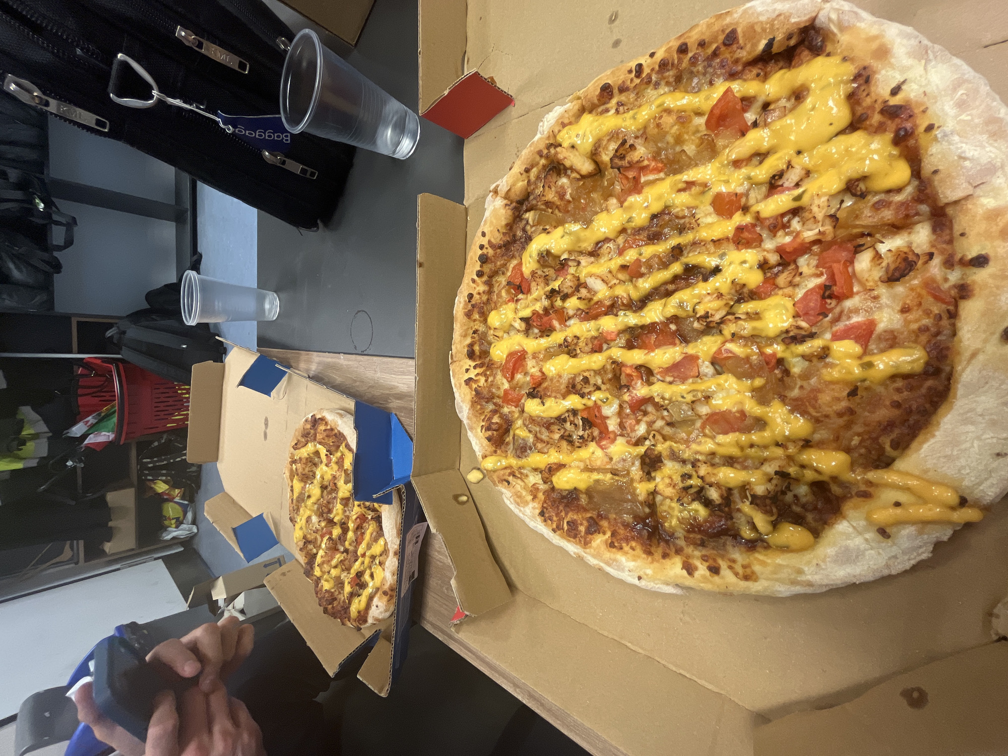 The image shows two pizzas in cardboard boxes, next to each other. A large pizza dominates the right side of the frame. The pizza has a thick crust, covered in toppings, including what appear to be tomato pieces and a yellow sauce drizzled across the pizza. The pizza sits inside a brown cardboard box. To the left, there is a smaller pizza in a similar box. The background of the image includes a black bag, a few empty plastic cups, and other miscellaneous items. In the lower left corner, a hand holding a dark electronic device (possibly a phone) is visible. The image does not contain enough specific information to identify the location in Sofia, Bulgaria.