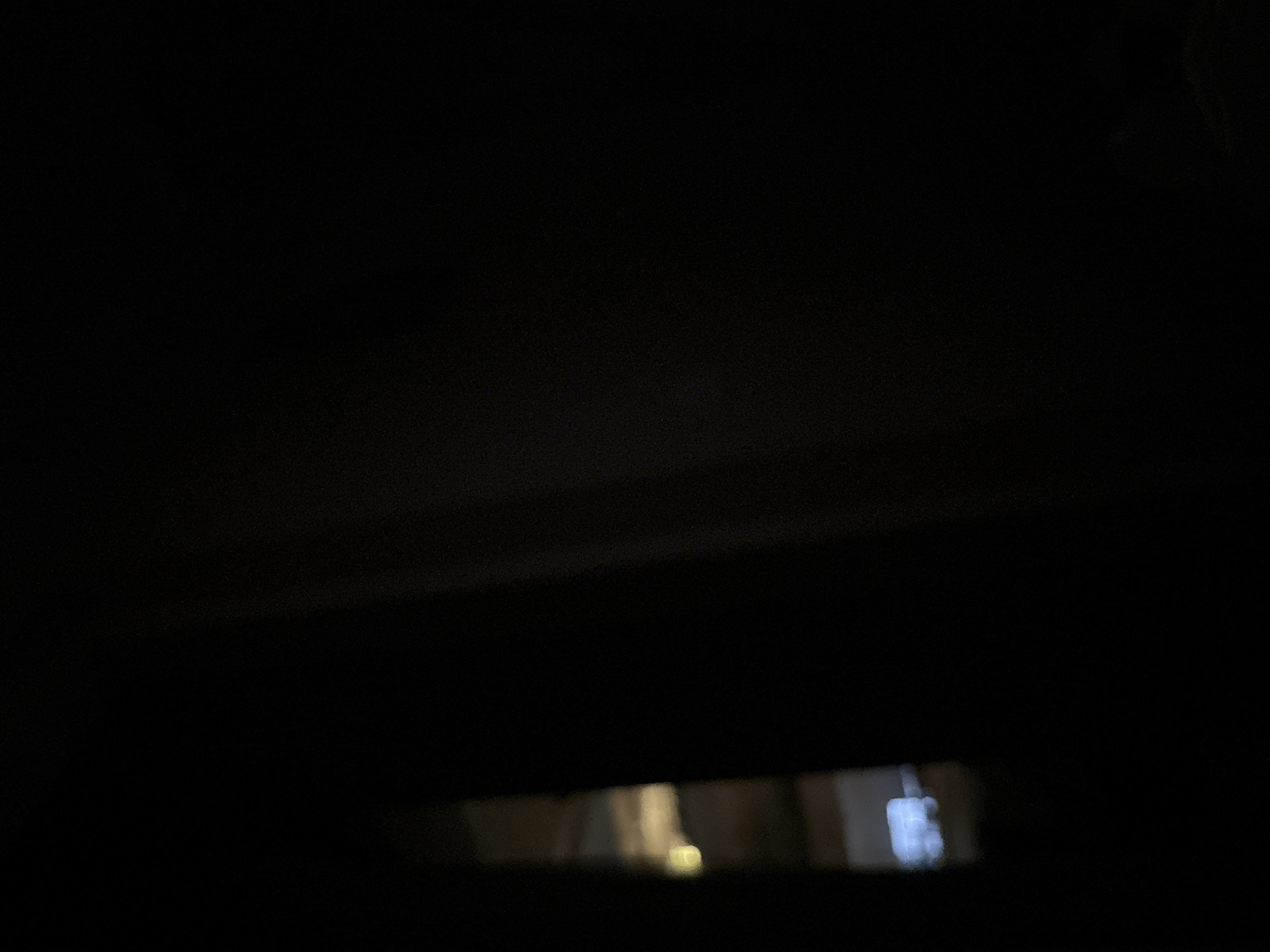 The image is predominantly dark with a sliver of light visible at the bottom. Through what appears to be a narrow opening, a glimpse of an interior space is visible. There is a suggestion of structure and possibly a light source within that space. The location in the city of Budva, Montenegro, cannot be determined from the image.