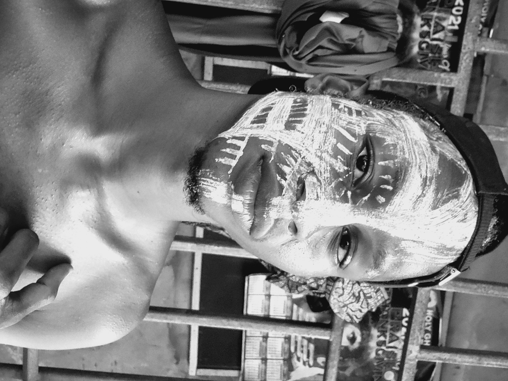 A black and white, vertically oriented portrait features a person, likely male, looking directly at the viewer. The subject's face is adorned with thick, white paint or chalk applied in distinct, abstract patterns, contrasting sharply with their dark skin. A short beard is visible, and the person wears a dark cap or headwear. Their right eye is prominently displayed, holding a direct and intense gaze.
The person is bare-chested, showing their shoulder, chest, and part of an arm. The skin appears textured and slightly moist, possibly indicating sweat. A patterned fabric is visible near their neck and shoulder.
In the background, a metal structure resembling bars or a fence is present. Behind this, various blurred elements, possibly signs or posters, are visible. Fragmented text can be deciphered, including "HOLY CH," "2021," and partially "ANKSGW," suggesting an outdoor or urban environment. The location is consistent with Onitsha, Nigeria. The monochrome processing emphasizes contours and textures, lending a stark and powerful quality to the image, which seems to capture a moment of cultural significance, performance, or personal expression.