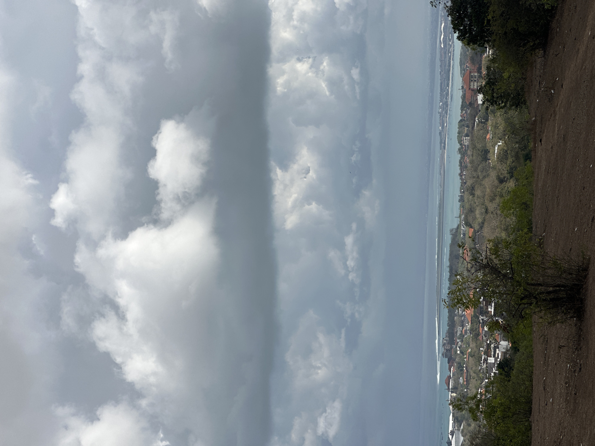 This is an aerial view of the city of Kuta Selatan, Indonesia, with a dramatic sky filled with gray and white clouds. In the foreground, there is a dirt hill with sparse vegetation. Below the hill, a densely populated area with white and red-roofed buildings stretches towards the coast. A body of turquoise water, likely the ocean, lies beyond the city, with a long sandbar or island in the distance. The overall atmosphere is overcast, suggesting potential rain.