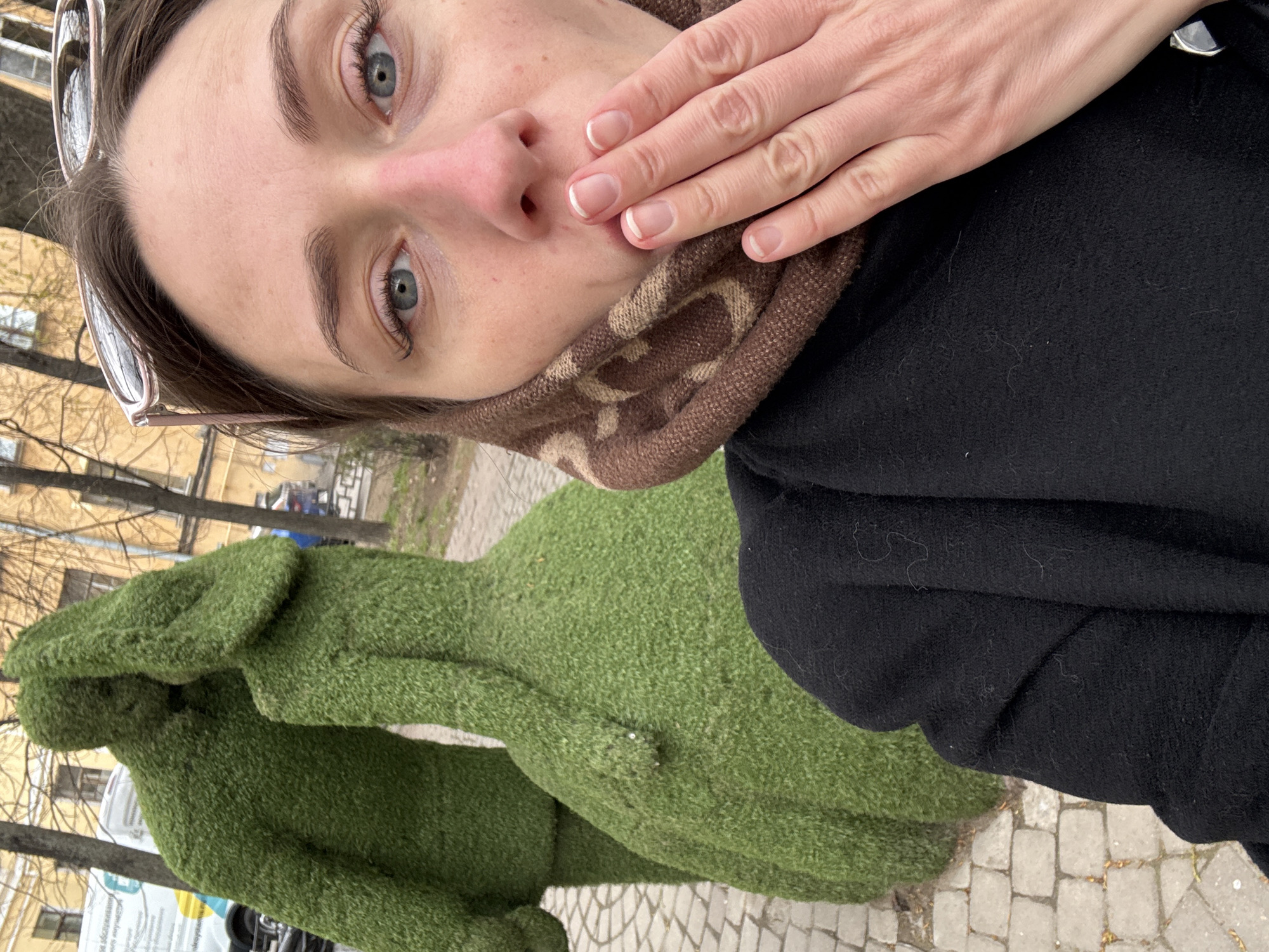 A selfie features a woman with light skin, blue eyes, and dark eyebrows, looking directly at the camera. Her dark hair is pulled back, and sunglasses rest on top of her head. Her left hand, adorned with a French manicure, covers her mouth in a gesture that could convey surprise, shyness, or contemplation. She is wearing a brown and beige patterned scarf or neck warmer and a black coat.
In the mid-ground, to her left, a large, vibrant green topiary sculpture, resembling a human figure (possibly a woman in a long dress), stands on a paved surface. The sculpture appears to be made of artificial grass or a similar textured material. The ground beneath is paved with light-colored cobblestones.
The background shows light yellow/beige buildings, some with visible windows, and several leafless trees, suggesting an outdoor urban or park setting. The sky is overcast, indicating daylight but no direct sunlight. In the distant background, faint, unreadable Cyrillic text is visible on a sign. The overall scene captures a moment outdoors, possibly on a cool day given the scarf and coat.