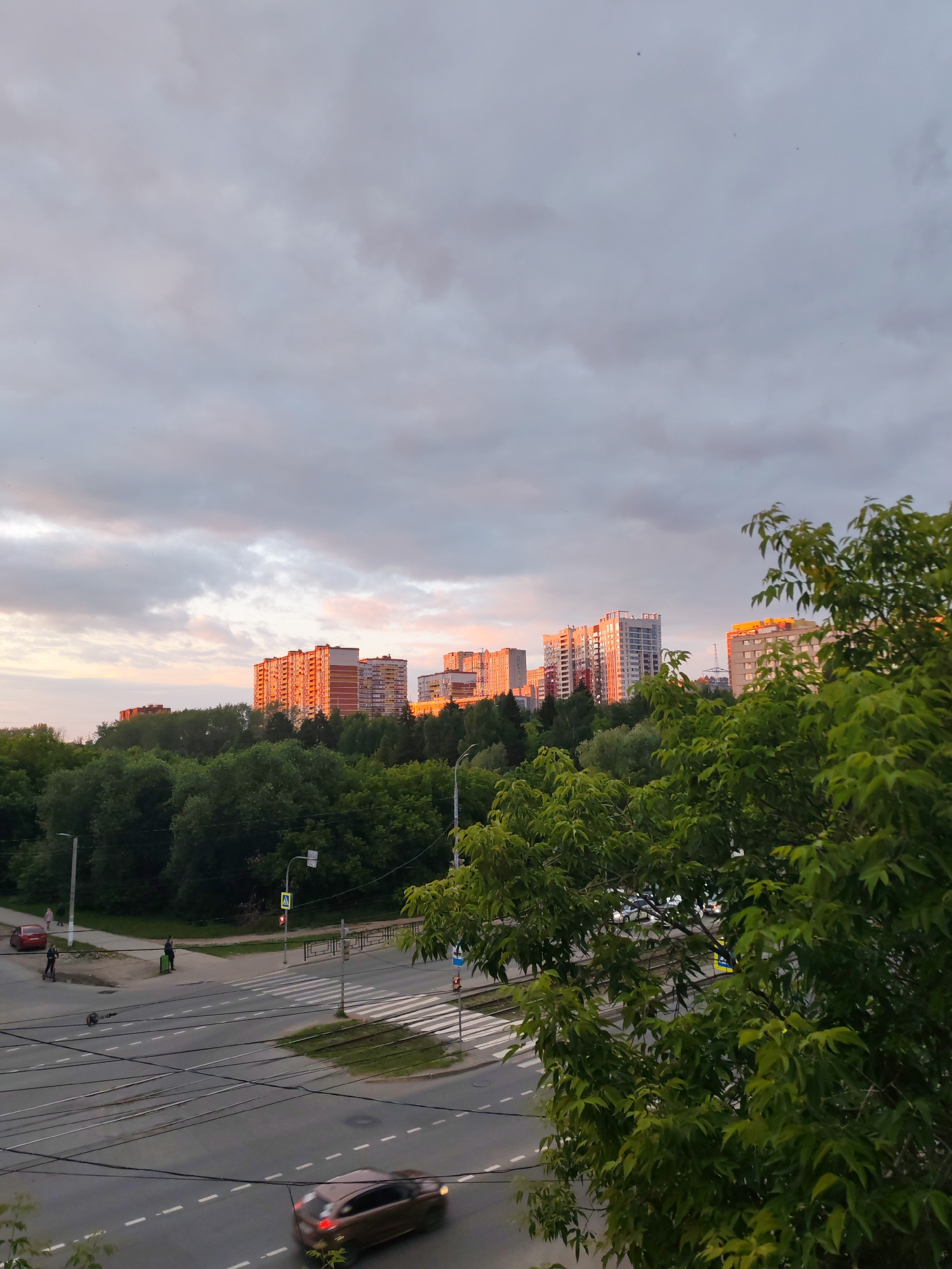 The image captures an urban scene in Izhevsk, Russia, during what appears to be dusk. In the foreground, lush green trees with dense foliage frame the view. Beyond the trees, a city street is visible, marked with white dashed lines and tram tracks. A brown car is seen in motion on the road, its brake lights illuminated. A pedestrian crossing is also present, with a traffic light displaying a red signal. A few people are observed on the sidewalk, and one individual is using a scooter. In the mid-ground, a line of trees forms a verdant border. Dominating the background are several multi-story residential buildings, their facades catching the warm, soft light of the setting sun, casting a reddish-orange hue on some of the structures. The sky above is overcast with clouds, exhibiting subtle shades of grey and hints of pink from the twilight.
