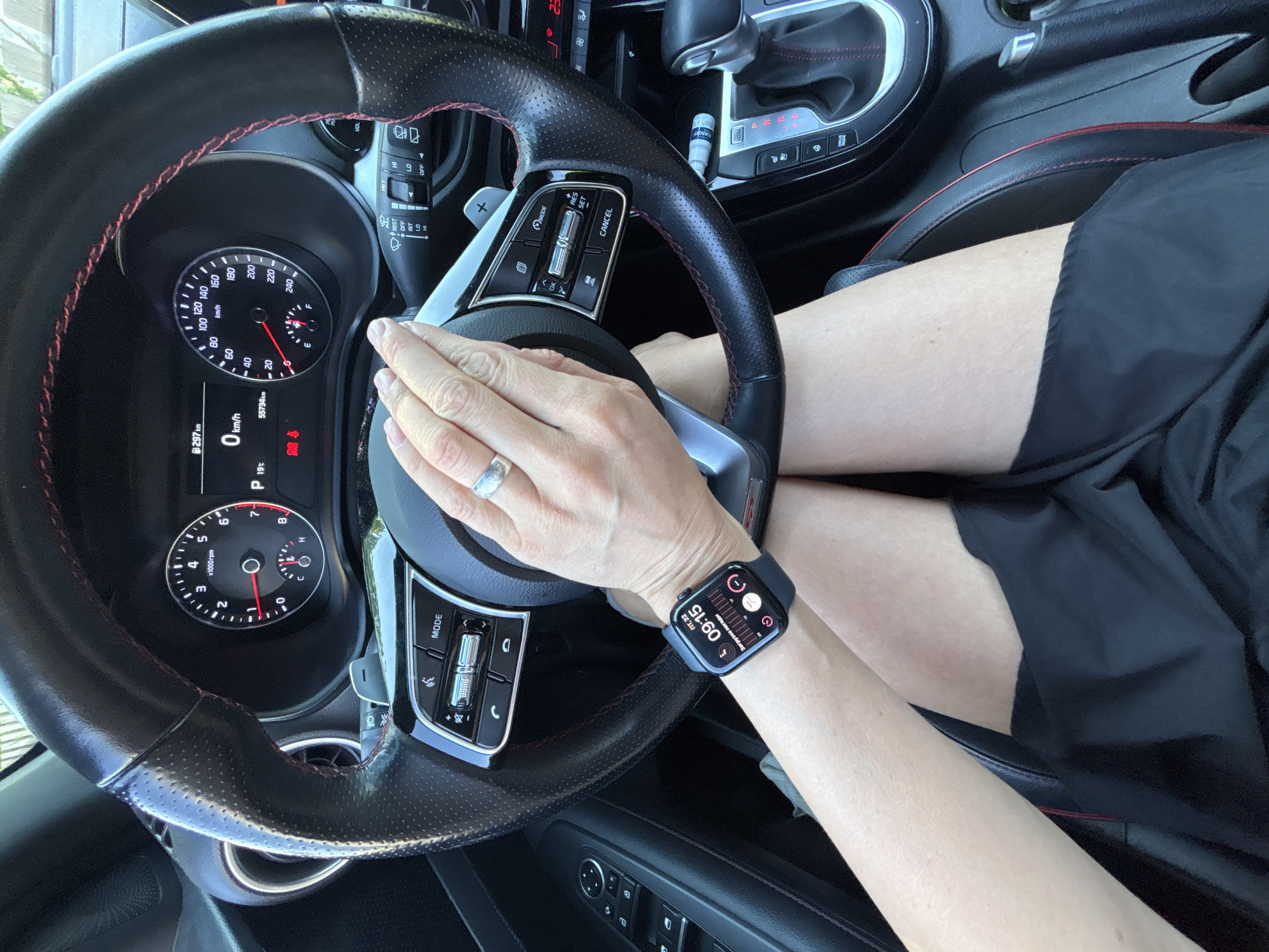 A person is driving a car. Their left hand is on the steering wheel with a silver ring on their finger. Their right wrist has a black Apple Watch displaying the time as 9:15. The car's speedometer reads 0 km/h and the odometer displays 55734 km. The image provides no information about the location in Chaikovsky, Russia.