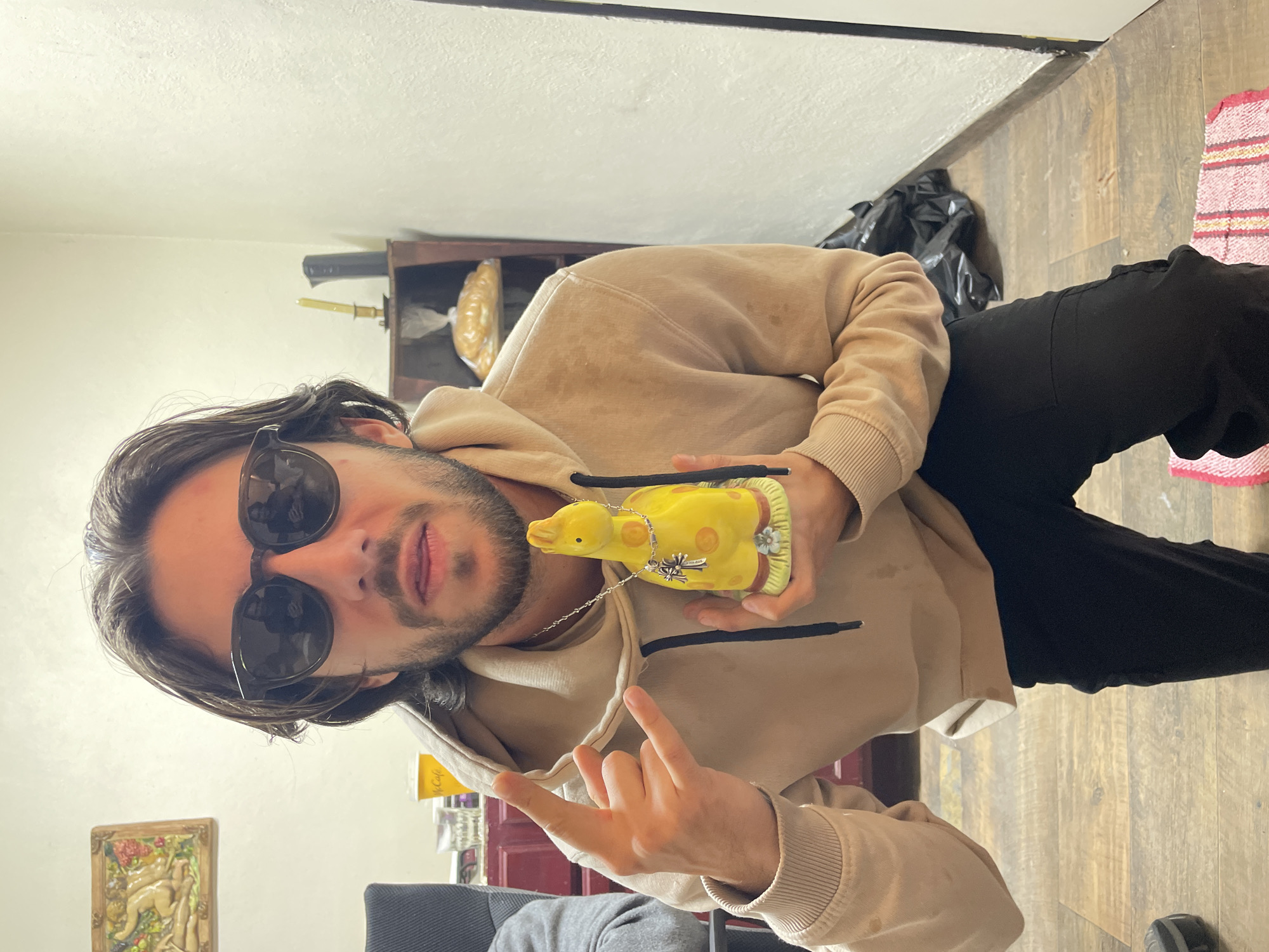 A man with long, dark hair and sunglasses is holding a yellow duck figurine with a silver chain around its neck. He is wearing a beige hoodie and black pants. His left hand is making a "rock on" gesture. The setting appears to be indoors, possibly a home. In the background, there is a wooden floor, a shelf with objects on it, a black plastic bag, and a rug. Based on the surroundings, the location could potentially be a residence or indoor space within Huixquilucan de Degollado, Mexico. The image composition suggests a casual, posed snapshot.