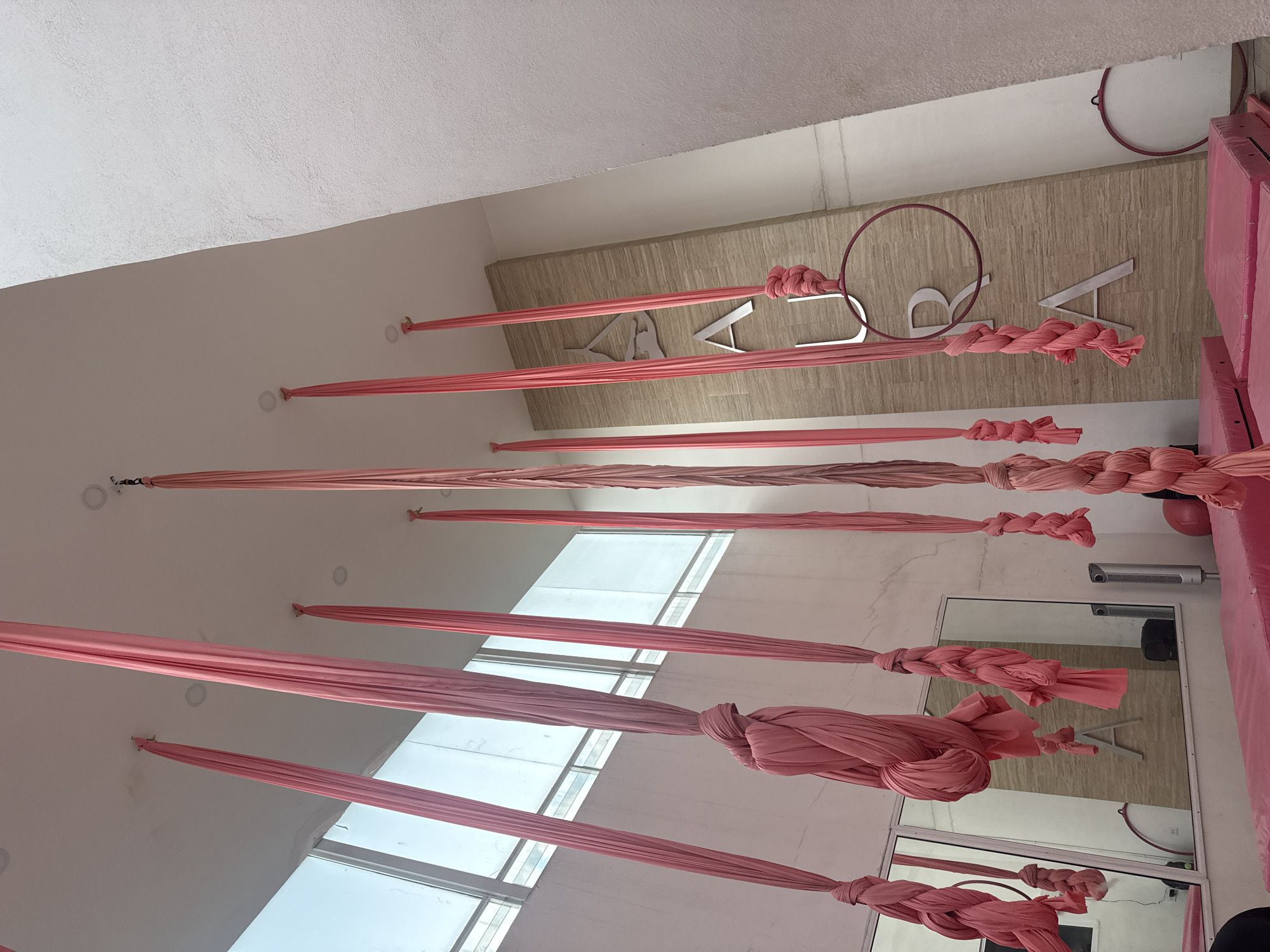 The image shows an interior space in San Andrés Cholula, Mexico, with pink aerial silks hanging from the ceiling. The silks are attached to the ceiling at regular intervals and are gathered and knotted at the ends. A wall is visible, decorated with a logo consisting of the word "VARDA" in silver letters and a pink hoop. Pink gym mats are leaning against the wall. A mirror reflecting part of the room is mounted on the wall. Light is coming in through windows. The scene is an aerial fitness or dance studio.