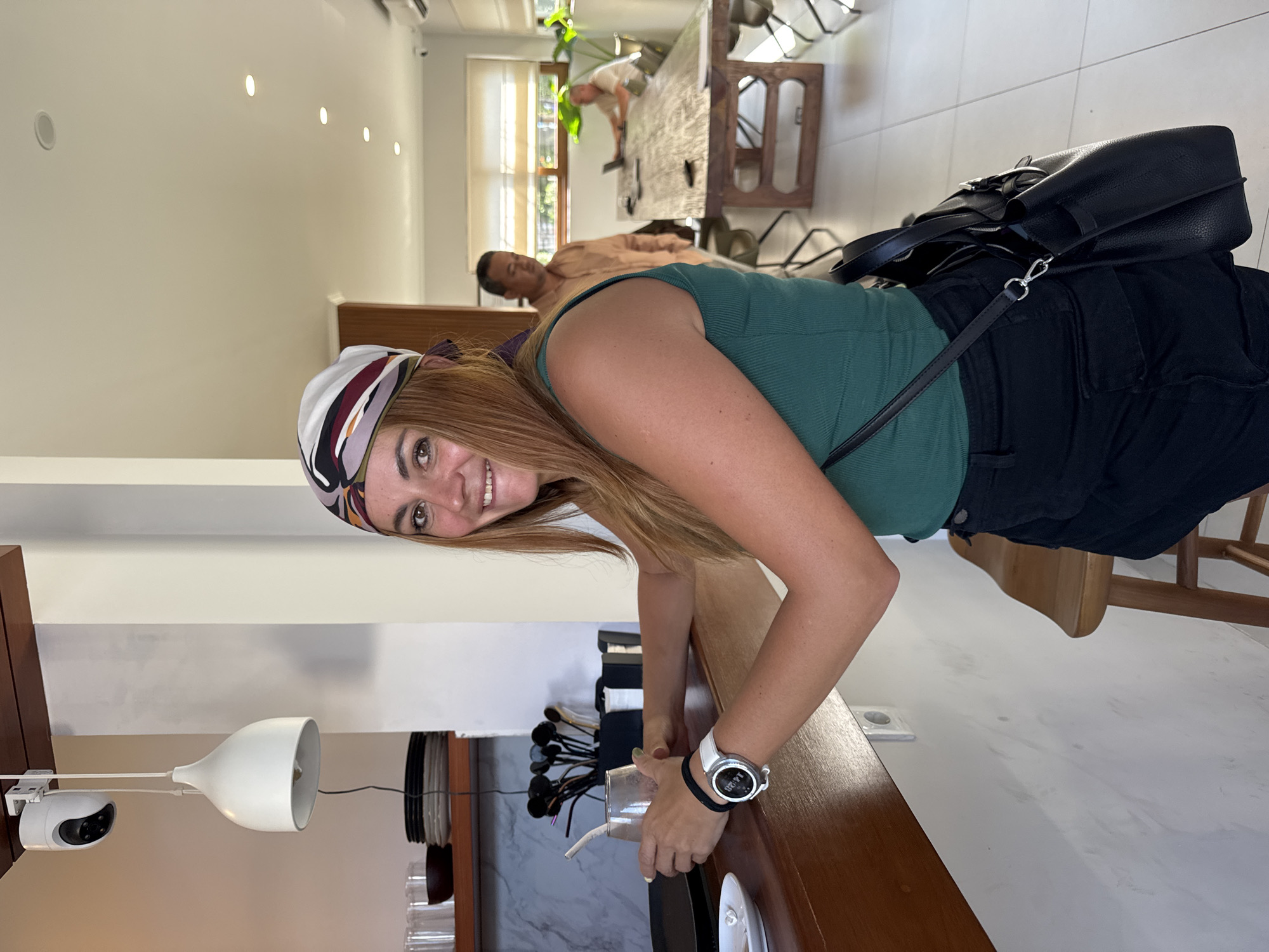 The image shows a woman with long brown hair, wearing a green tank top, black shorts, a patterned headscarf, and a watch, leaning over a wooden counter. She is holding a glass with a straw in it. On the counter are plates, cutlery, and a lamp. In the background, there is a dining area with people, wooden tables, and chairs. The location is Denpasar, Indonesia.