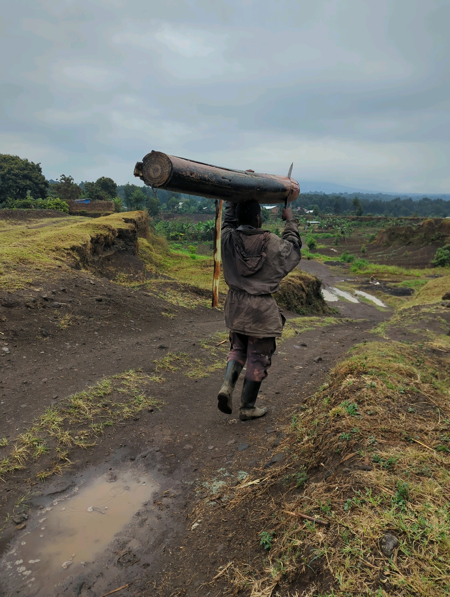 A person walks away from the camera carrying a large, cylindrical object horizontally on their shoulders. The person is wearing a dark hooded jacket, patterned trousers, and tall black rubber boots. They are walking on a dirt path with patches of grass and muddy puddles. The path is on the edge of a steep, eroded embankment covered in dry grass. In the background, there is a rural landscape with trees, fields, and some buildings. The sky is overcast and gray. The scene appears to be located in the Musanze District of Rwanda.
