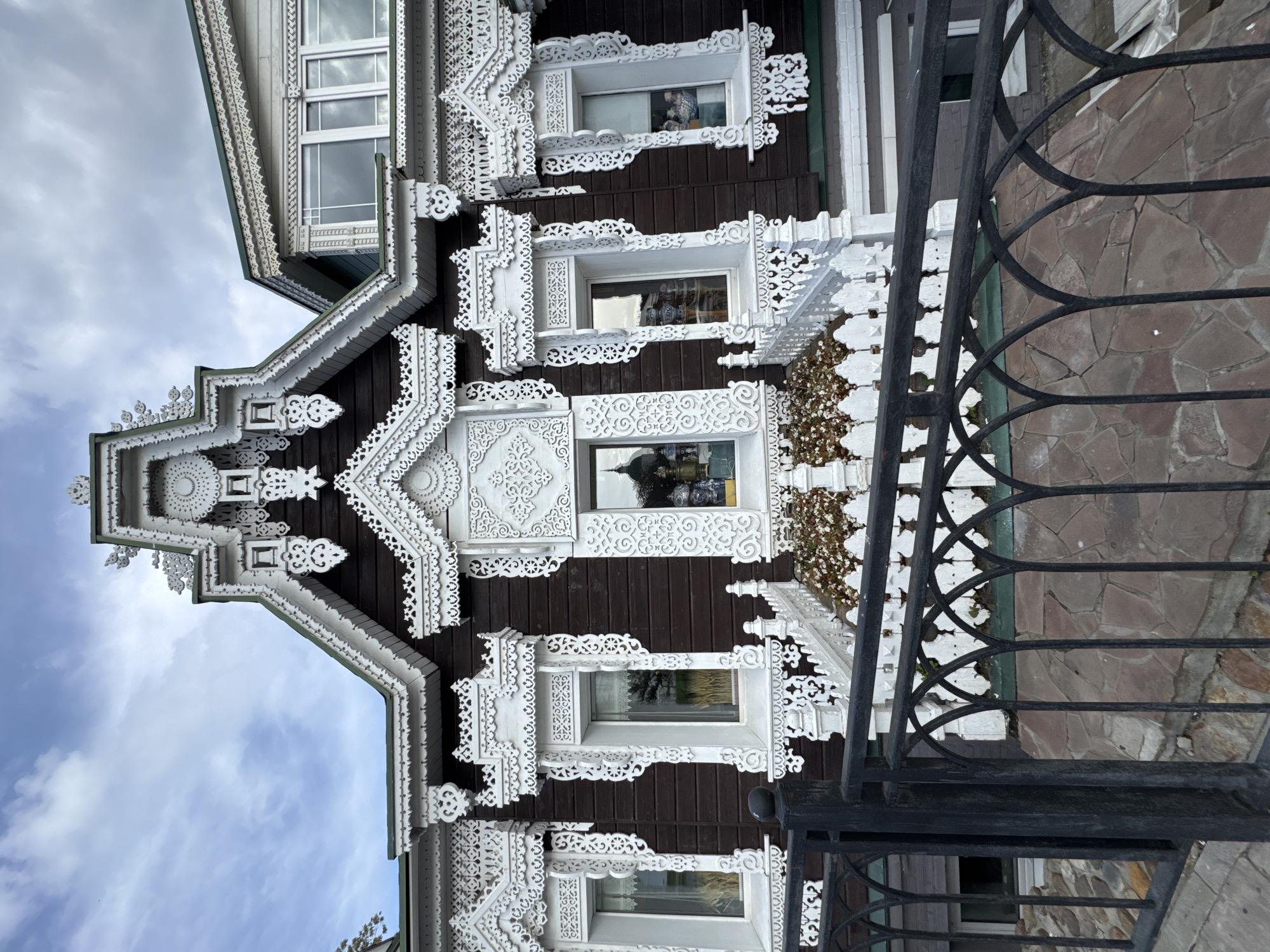 The image shows a close-up view of the facade of a wooden house in Sergiyev Posad, Russia, known for its ornate "terem" style architecture. The house has a dark brown wooden exterior with intricate white wooden carvings adorning the eaves, window frames, and gables. These carvings feature detailed patterns of geometric shapes, floral motifs, and lace-like designs, characteristic of traditional Russian wooden architecture. Several windows are visible, each framed by these elaborate white elements. A white railing and planters with small flowers are positioned in front of the windows, leading up to a central entrance or bay window. A black metal fence with arched elements is in the foreground, partially obscuring the lower part of the house and the stone pathway behind it. The sky above is partly cloudy. The architectural style and the surrounding elements suggest this is likely a historical building, possibly a museum or a traditional residence, located within the city of Sergiyev Posad.