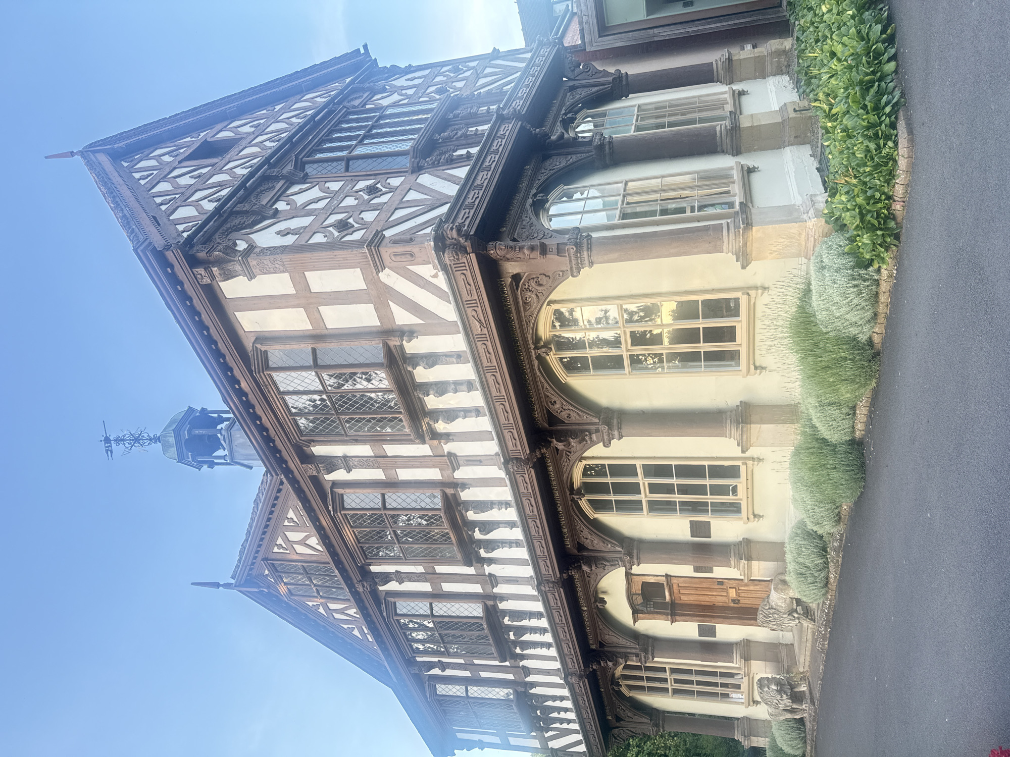 The image shows a detailed, three-story Tudor-style building. The facade is characterized by its distinctive half-timbered construction, with dark wooden beams set against a light-colored plaster. The upper floors feature numerous diamond-paned windows, many of which are in bay window configurations. Intricate wooden carvings adorn the eaves and decorative elements are present along the facade. The ground floor has a pale yellow stucco finish and is punctuated by arched windows with similar diamond panes. A wooden door is centrally located on the ground floor. Flanking the entrance steps are two stone lion statues. In front of the building, there is a paved pathway or road and a garden with low, bushy plants and some taller greenery. The sky is clear and blue, suggesting daylight. The building appears to be situated in a well-maintained outdoor setting. The architectural style and potential historical significance suggest this could be a public building or a notable residence. Based on architectural clues, it is likely the Grangefield Grange in Leominster, United Kingdom, which is known for its historical Tudor architecture.