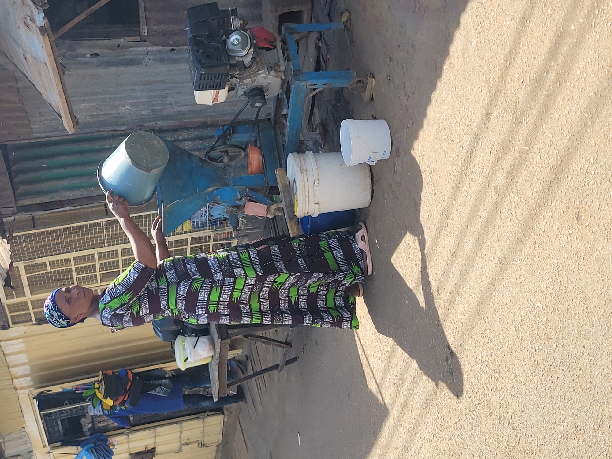 An outdoor scene in Jos, Nigeria, captures a woman engaged in processing goods with a grinding machine under bright sunlight. The woman, dressed in a vibrant green, black, and white patterned gown and a blue headwrap, is positioned next to a robust blue grinding machine. She holds a blue bucket, appearing to either pour ingredients into the machine or prepare to do so. The grinding machine features a visible engine, with a faint "HONDA" logo discernible on its side, and various pulleys and belts.
Surrounding the machine are several plastic buckets, mostly white and one blue, likely used for holding raw materials or processed products. The ground beneath the machine and around the woman is a sandy, dusty surface with fine grain particles scattered across it, indicating the nature of the grinding operation. Strong, clear shadows are cast by the woman, the machine, and other objects, suggesting it is midday or early afternoon. In the background, structures made of corrugated metal sheets are visible, characteristic of market stalls or small workshops. The scene depicts a moment of daily labor and commerce in a local setting.