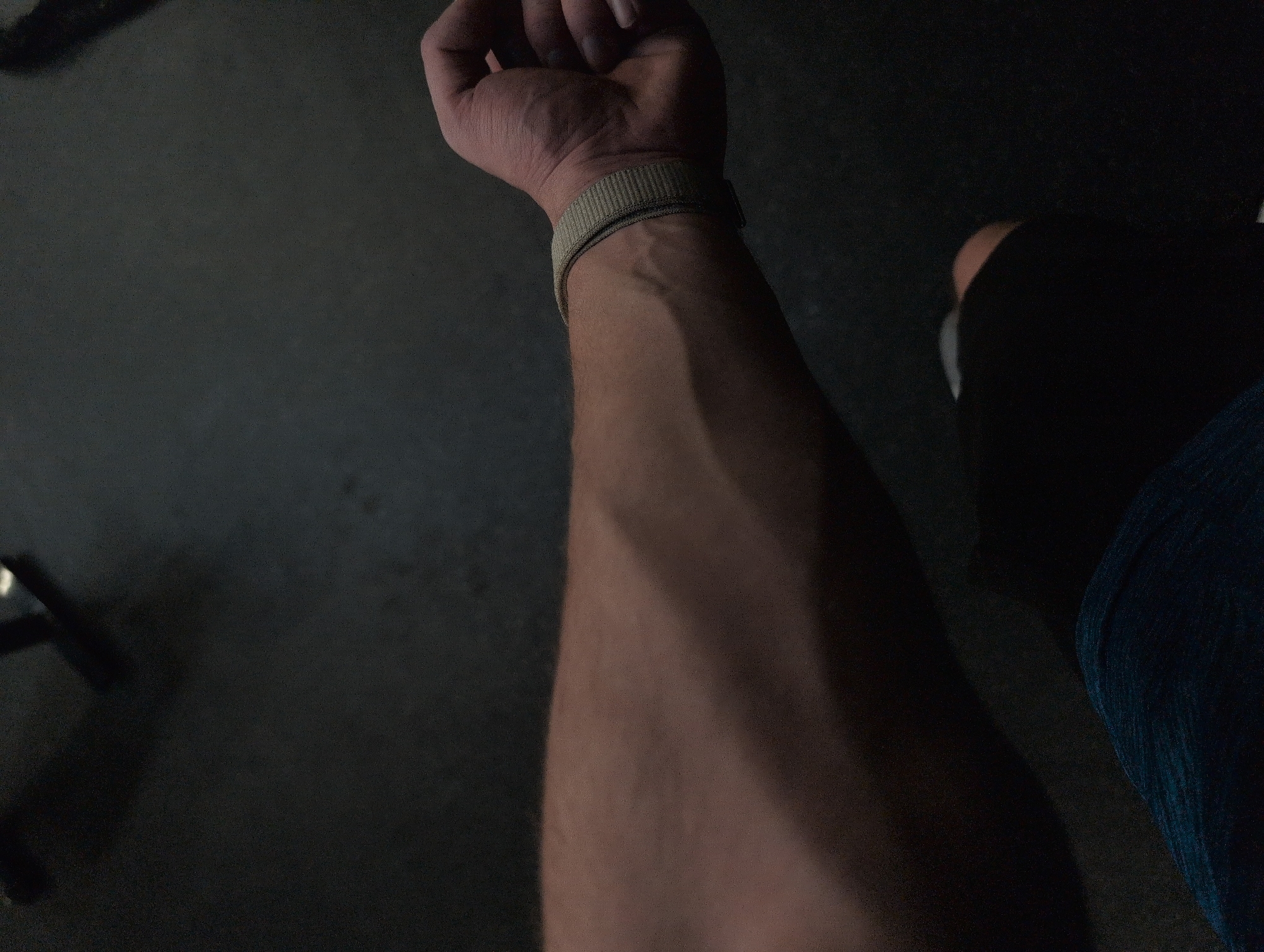 The image presents a close-up, high-angle view of a person's left forearm and hand in a dimly lit environment. The arm extends from the upper left corner towards the bottom right, with prominent veins visible on the forearm and the back of the hand, suggesting a lean physique or recent physical exertion. A light-colored, possibly beige or olive green, fabric wristband is worn on the wrist, securing around the lower part of the forearm. The hand's fingers are slightly curled.
The background is predominantly dark and out of focus, appearing to be a floor or ground surface with some subtle texture. In the bottom right quadrant of the image, a small portion of what appears to be a person's leg or thigh is visible, clad in dark fabric that could be blue jeans or athletic wear.
The scene is characterized by low, ambient lighting, which creates deep shadows and obscures finer details in the surroundings. There are no discernible specific environmental cues to pinpoint an exact location within Mexico City, Mexico, nor are there any indications of weather or time of day beyond the general low light conditions. No text is visible in the image, and no specific activity or emotion is clearly conveyed.