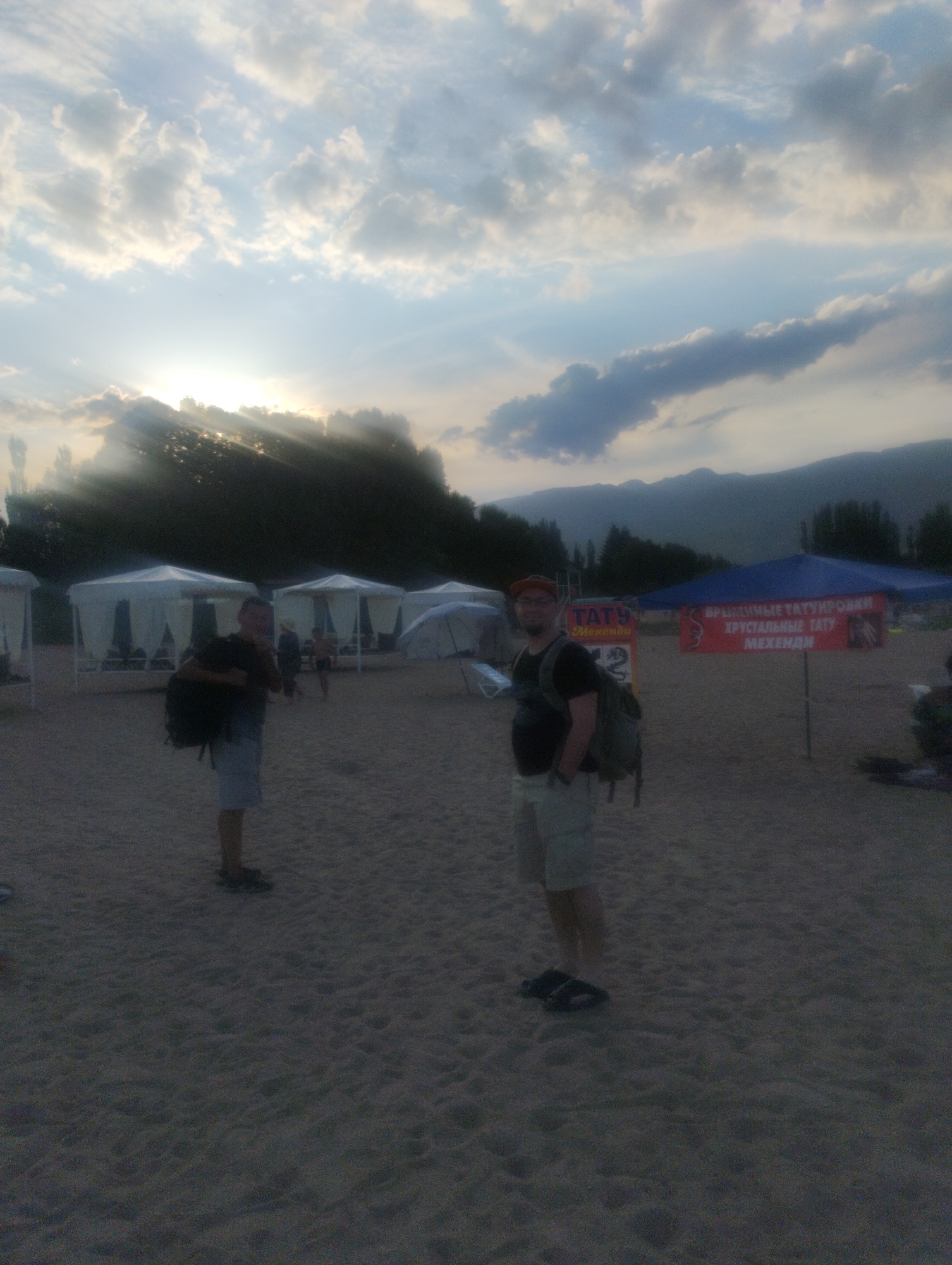 A medium shot shows two men standing on a sandy beach in Cholpon-Ata, Kyrgyzstan. The man on the left, facing away from the camera, wears a dark t-shirt, light shorts, and a backpack. The man on the right, facing the camera, wears a dark t-shirt, light shorts, a baseball cap, and a backpack. He has his right hand in his pocket. In the background, white tents with open sides are set up on the beach. A blue canopy with red banners advertising temporary tattoos is visible to the right of the man on the right. Mountains are visible in the far distance. The sky is cloudy with the sun setting, casting rays of light through the clouds. The overall scene depicts a beach resort area during a sunset.