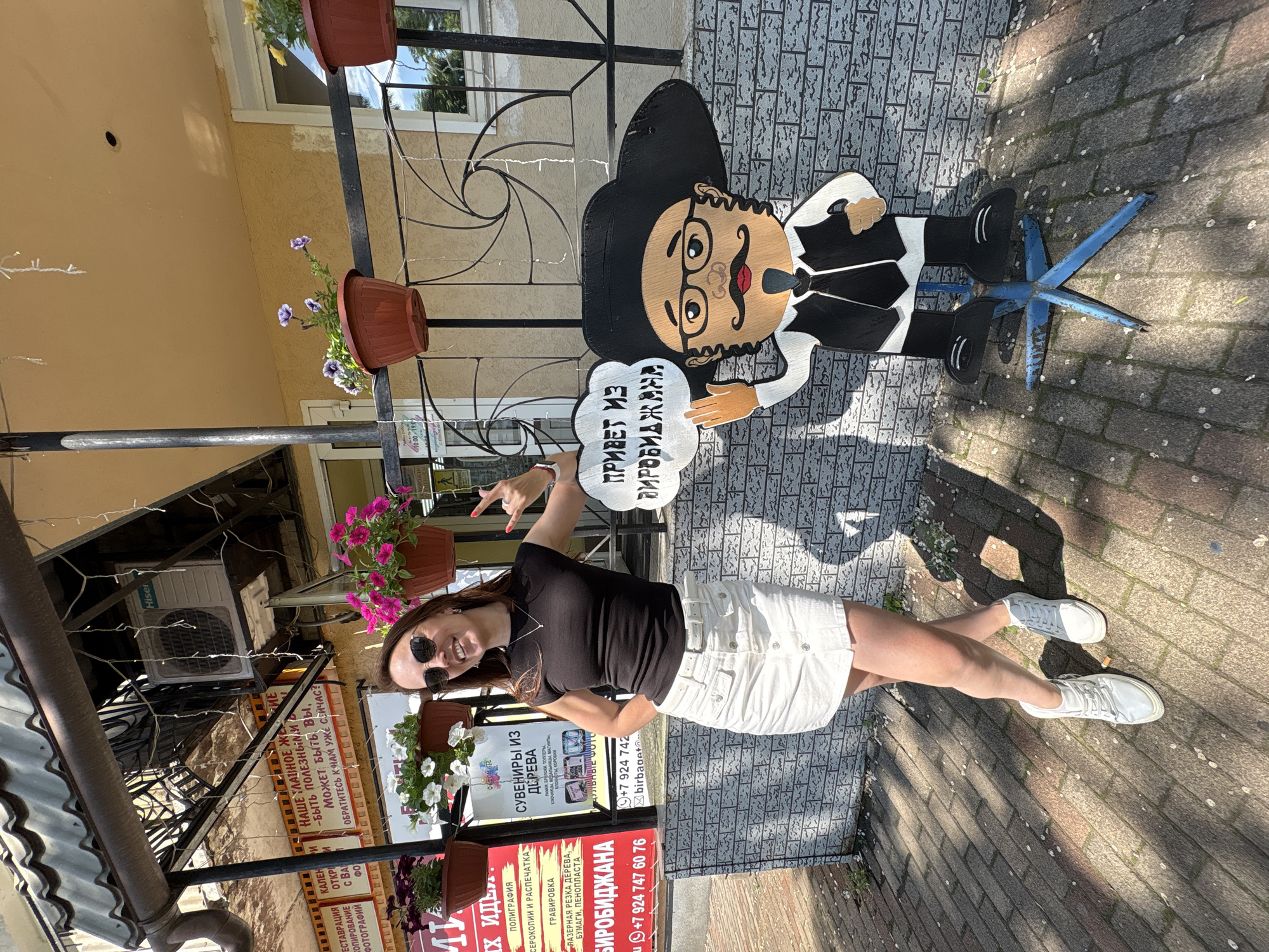 A woman with dark hair and sunglasses stands on a paved sidewalk in front of a building with a covered porch. She is wearing a black sleeveless top, a white denim skirt, and white sneakers. She has her left hand on her hip and is making a rock-on hand gesture with her right hand. Next to her is a wooden cutout of a man with a large black hat, glasses, a mustache, and a beard, wearing a black vest and tie. The cutout holds a speech bubble that reads "ПРИВЕТ ИЗ БИРОБИДЖАНА," which translates to "Greetings from Birobidzhan." Signs and potted flowers are visible on the building behind them. The scene is illuminated by sunlight, casting shadows on the ground. The building appears to be a shop or business, with advertising visible on the signs. The overall impression is a tourist snapshot taken in Birobidzhan, Russia.