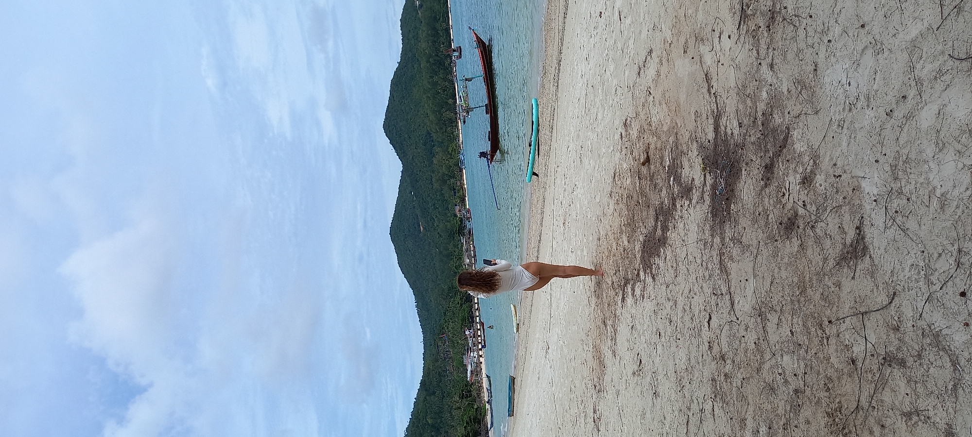 The image shows a woman with curly brown hair standing on a beach in Baan Chaloklum, Thailand. She is wearing a white long-sleeved outfit and holding a mobile phone. The beach is sandy with scattered debris. In the background, there is a green-covered mountain, the ocean with a longtail boat and a paddle board, and the sky with light clouds.
