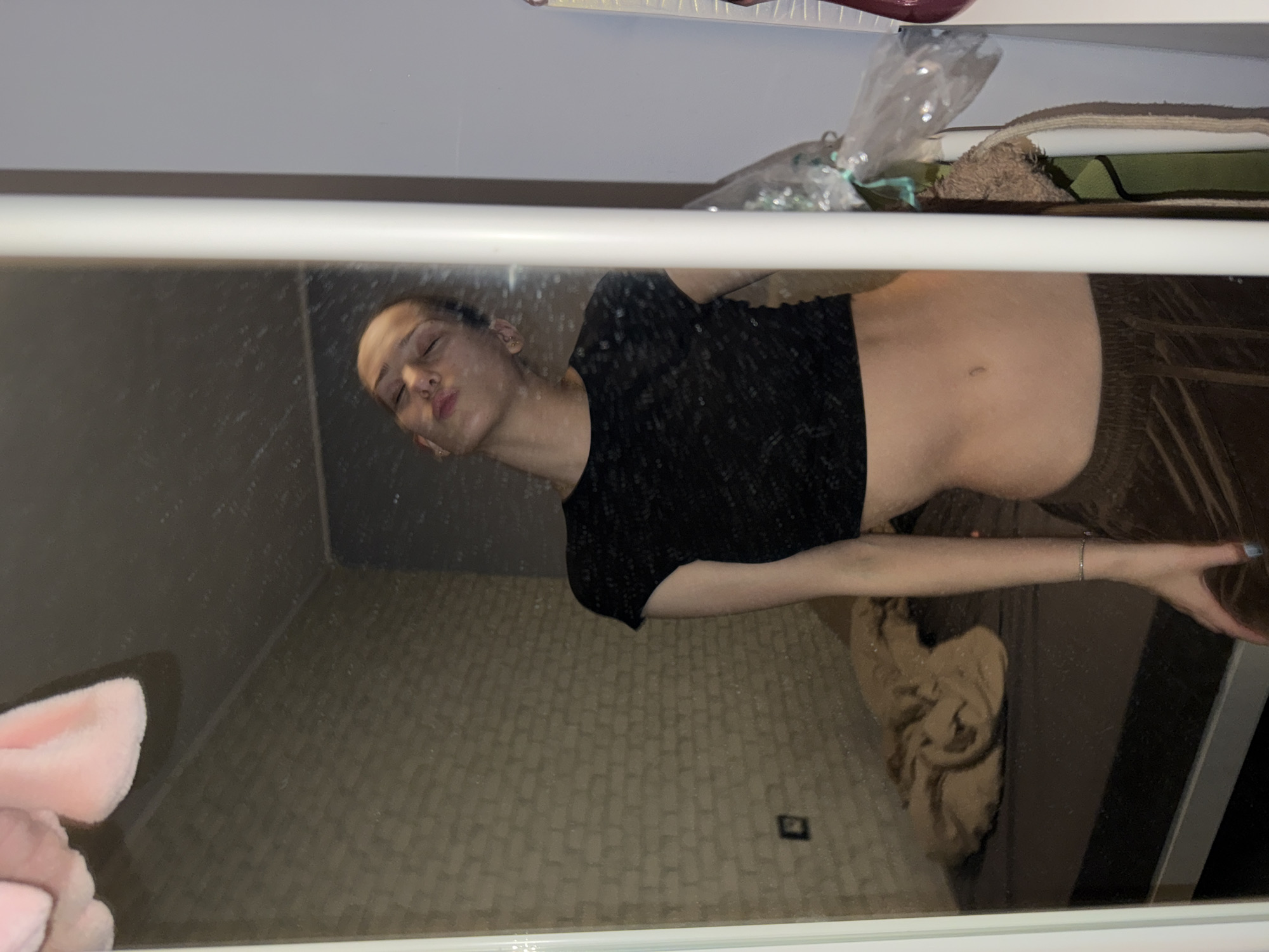 The image shows a reflection of a person in a mirror or a shiny surface. The person has short dark hair, wearing a black top, and brown pants. Their face is turned towards the left with their lips puckered. The background reflected in the mirror shows a tiled wall and a towel on the floor. Above the mirror, there are various items including a plastic bag and a towel. The image location is not discernible and it is not possible to identify the location as Sofia, Bulgaria.