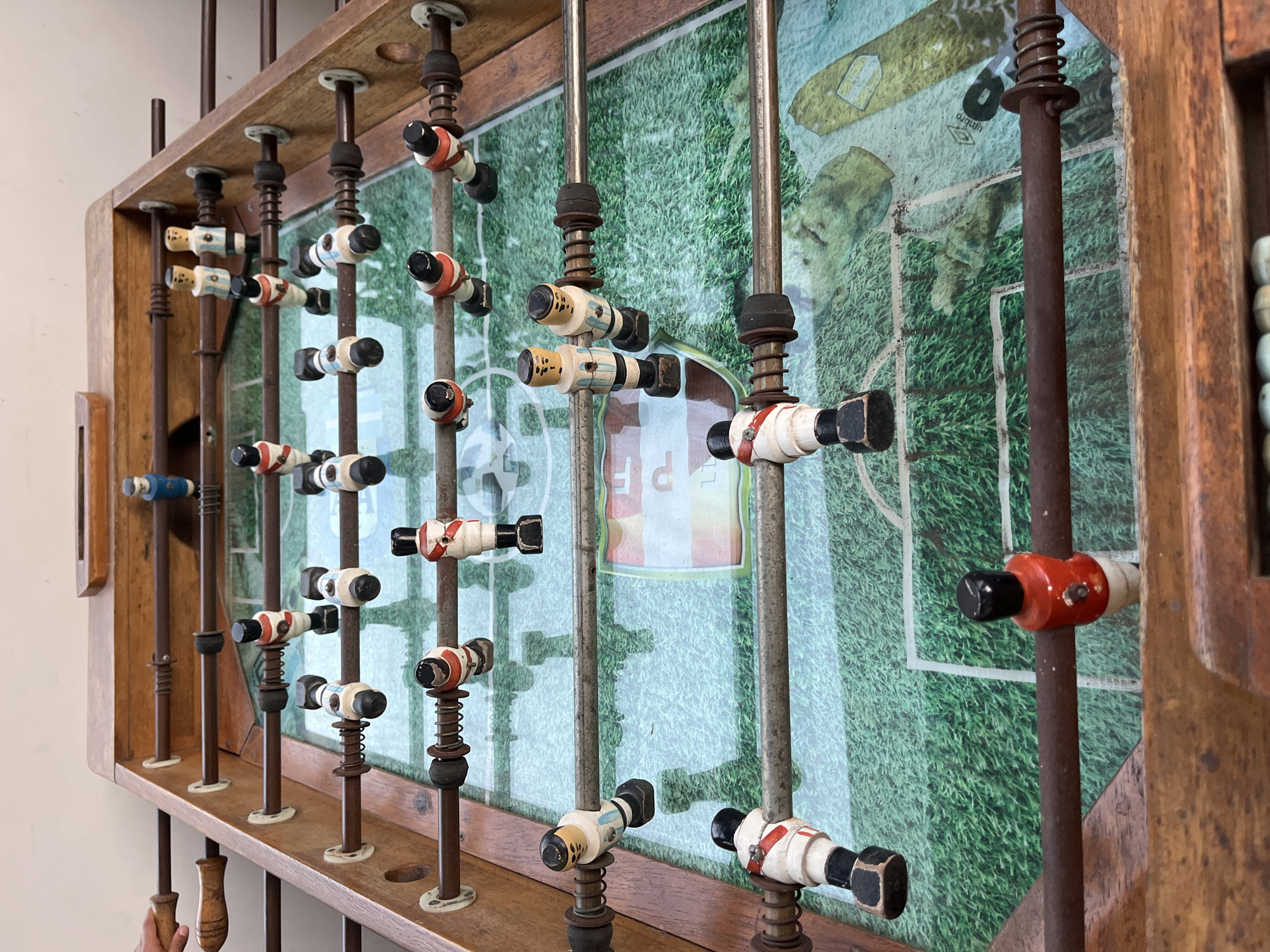 A slightly high-angle, indoor close-up shows a vintage foosball table in a wooden frame. The table features metal rods with small, multi-colored plastic players attached. The players are primarily white with black ends and some red or blue markings. The playing surface is visible in the background, depicting a green soccer field with faint logos and text. The rods extend through holes in the wooden frame, and some are equipped with springs and metal fittings. The overall condition of the table suggests age and use. It is not confirmed that the photo was taken in Pacarán, Peru.
