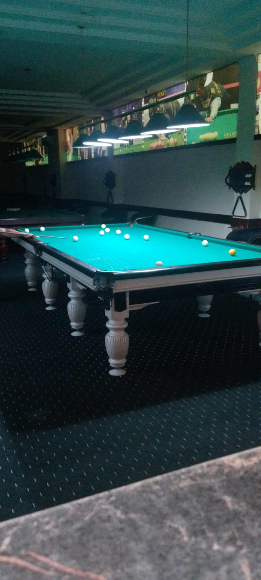 The image shows an indoor billiards hall in Angren, Uzbekistan, with two pool tables visible. The main focus is on a pool table in the foreground, covered in green felt, with several white balls scattered across its surface. One yellow ball is also present. A pool cue is resting on the table, suggesting an active game. The table has ornate white legs. In the background, a second pool table is visible, partially obscured. The walls are decorated with posters, one of which prominently displays the word "START". Overhead, several industrial-looking light fixtures illuminate the space. The floor is covered in dark carpeting with a repeating pattern. The overall ambiance suggests a recreational or entertainment venue.