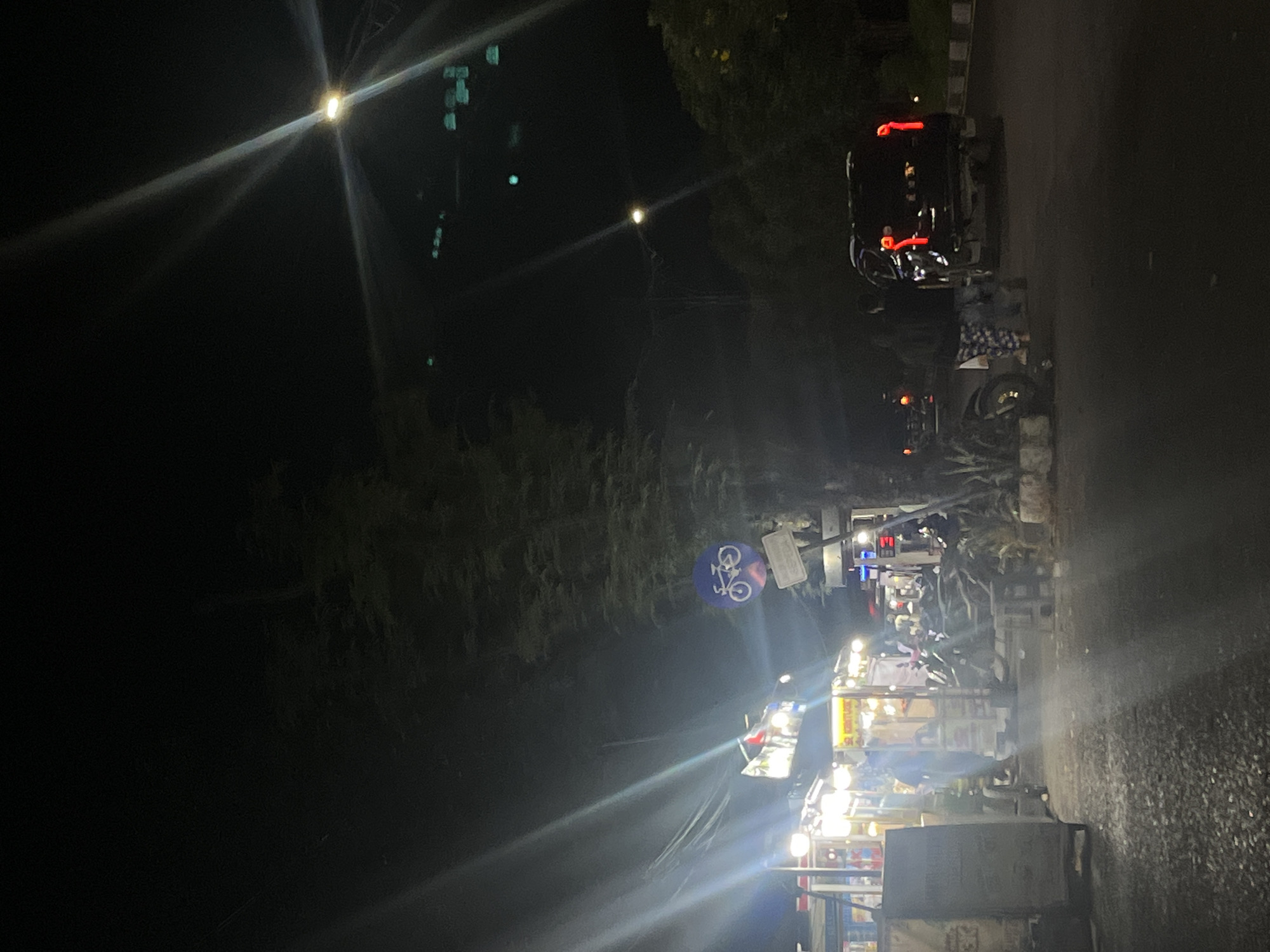 This is a nighttime street scene in Banda Aceh, Indonesia, with artificial lighting from streetlights and vendor stalls. In the foreground, the asphalt road surface is visible, with some debris. To the left, a street food stall is illuminated by bright lights, with several motorcycles parked nearby. One person is partially visible behind the stall. Further down the street, a dark-colored SUV is parked, with its brake lights illuminated. Two individuals are standing near the SUV, one facing away from the camera and the other partially obscured. A blue traffic sign with a white bicycle symbol is mounted on a pole to the right of the food stall. Trees and foliage are present in the background, and the sky is dark, with visible light flares from artificial light sources.