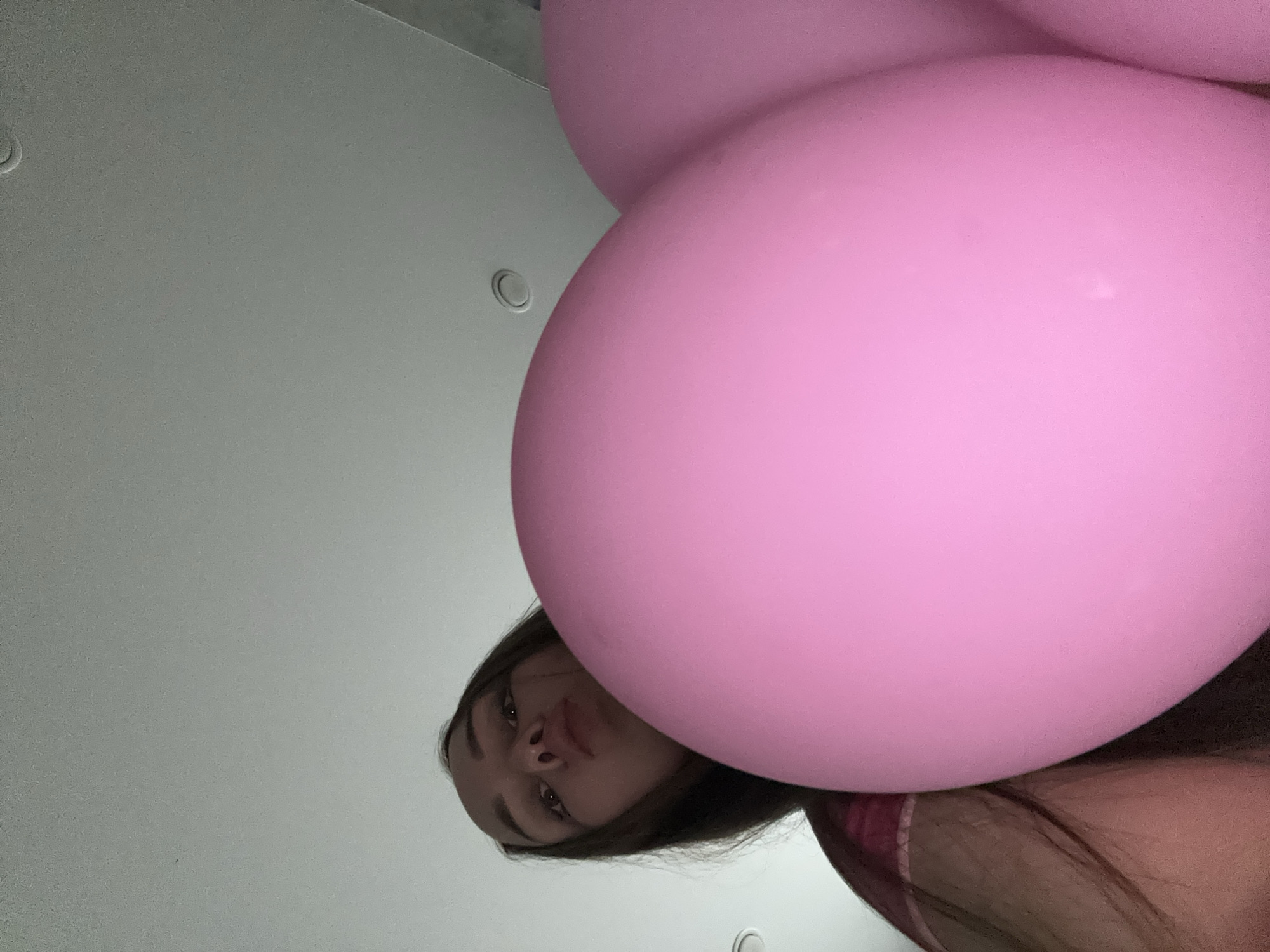 A low-angle shot shows a young woman partially obscured by large, pink balloons. The background is a plain, white wall. The location is unknown and there is no information to indicate the city of Chaikovsky, Russia.
