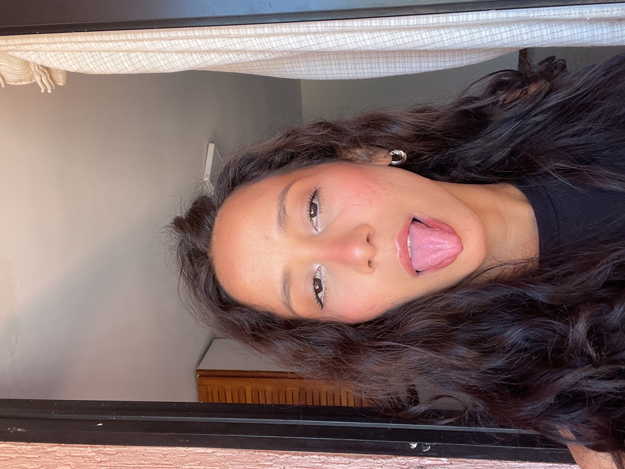 A medium shot shows a young woman with dark, curly hair looking directly at the camera with her tongue sticking out. She is wearing a black top and silver hoop earrings. The background consists of a textured pink wall on the left, a wooden cabinet and a white curtain on the right, suggesting an indoor setting, possibly an apartment in Porlamar, Venezuela. The lighting suggests it is daytime, with natural light coming from a window or opening to the right.