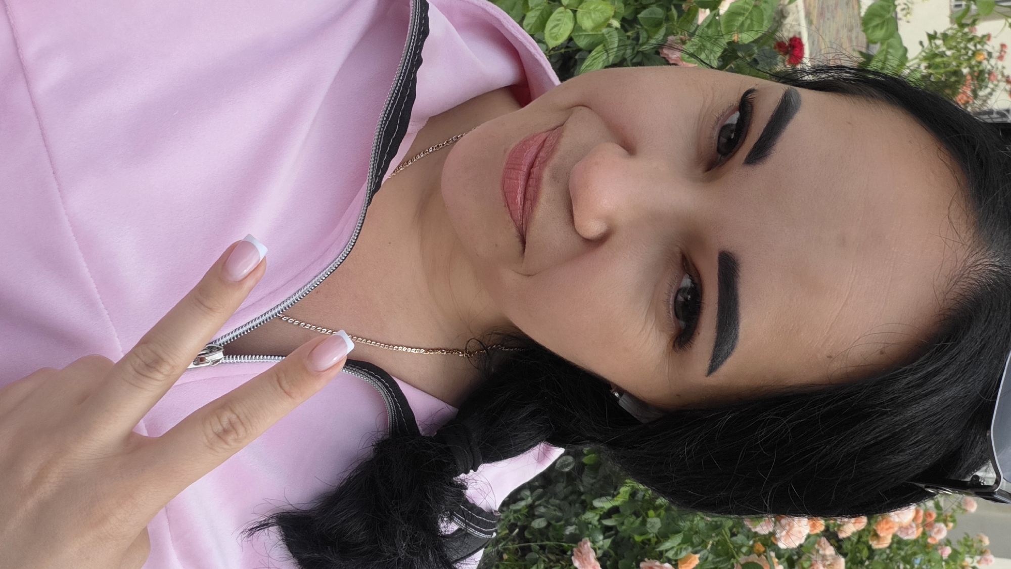 A woman with dark hair in a braid, wearing a pink jacket and a gold chain necklace, is shown from the chest up, outdoors. She is holding up two fingers in a peace sign gesture. Roses and greenery are visible in the background, suggesting a garden or park setting.