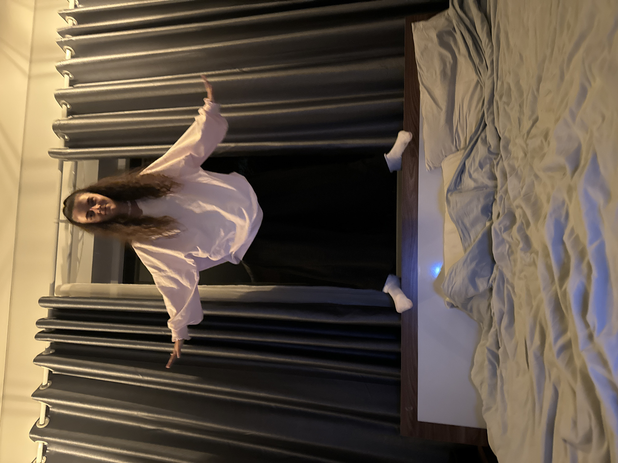 A person with long, curly brown hair, wearing a light pink sweatshirt and black pants, is lying on a bed. They are positioned horizontally in the frame, with their head towards the left and feet towards the right. Both arms are extended outward. The bed is made with light-colored sheets and pillows. Behind the person is a wall covered with grey curtains. There is no visible evidence that this image was taken in Da Lat, Vietnam.
