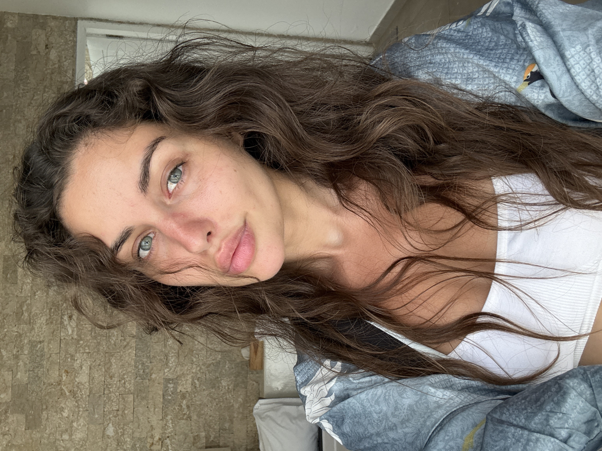 A close-up, rotated portrait of a woman with long, wavy brown hair. She has light skin, light blue eyes, and natural-looking lips. She is wearing a white top, partially visible, and a blue patterned jacket. The background consists of a stone wall texture on one side and a blurred view of a white door or window frame. The location is unknown, Indonesia.