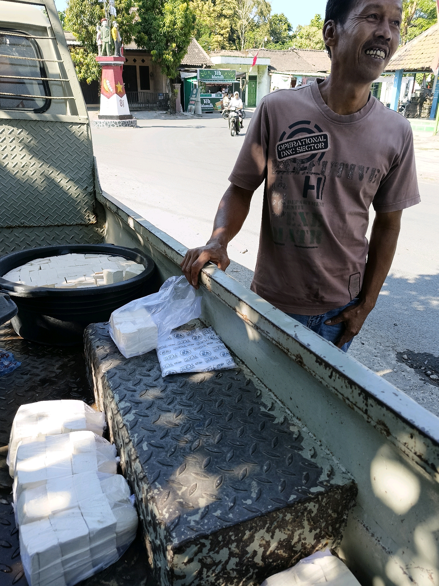 In Sukorini, Indonesia, a man stands in the back of a pickup truck filled with tofu. The man, wearing a brown t-shirt and blue jeans, is smiling and appears to be a vendor. The truck bed is partially covered with a diamond plate metal surface. In the foreground, several packages of tofu are visible, some in plastic bags and some in a large black tub filled with water. In the background, a street scene unfolds with buildings, trees, and a monument. The monument, a red and white structure with figures on top, appears to be a local landmark. The overall scene depicts a typical day in the city, with commerce and local culture evident.