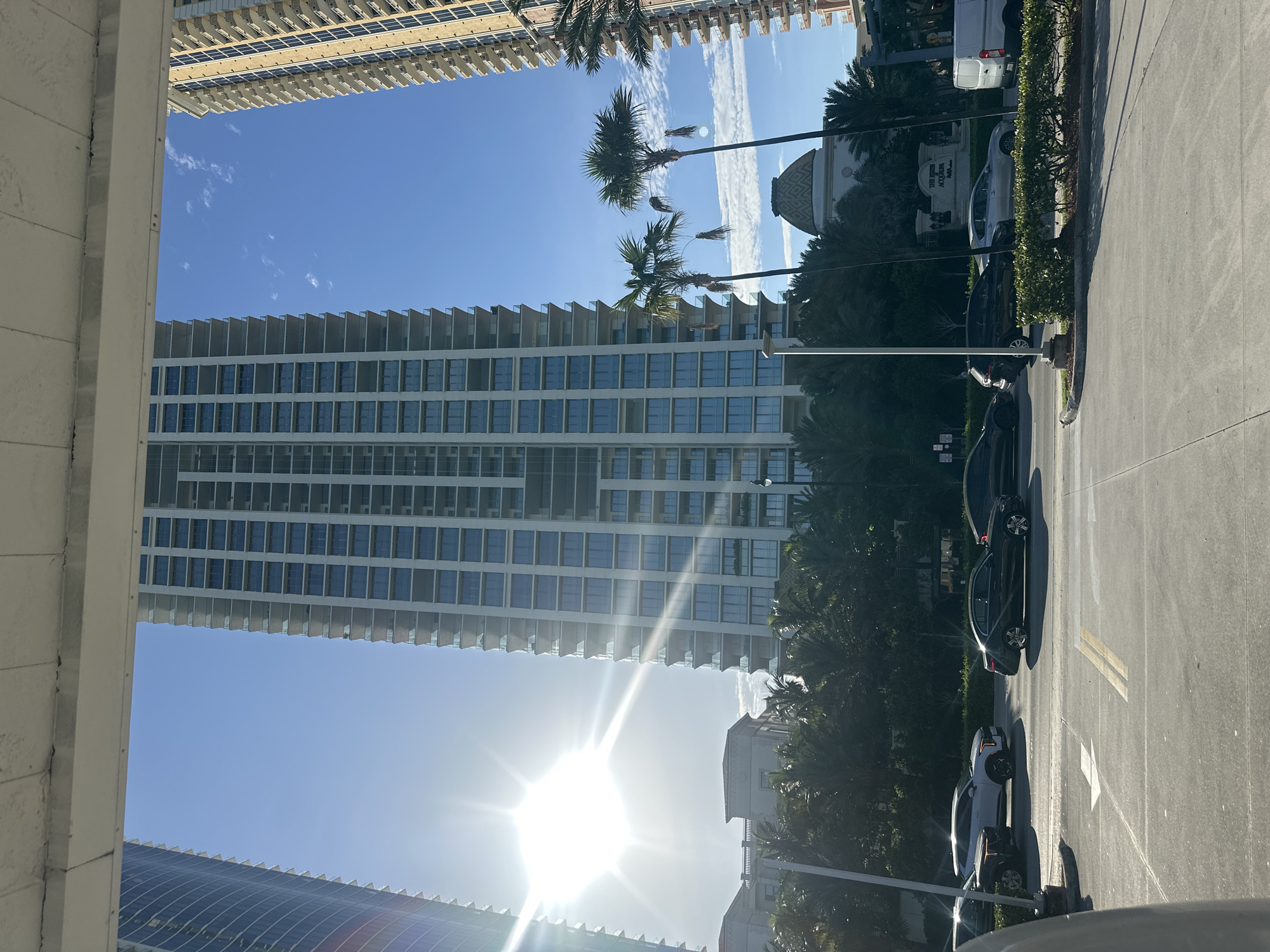 The image captures a sun-drenched street scene in Sunny Isles Beach, Florida. Dominating the frame are several tall, modern residential buildings, with a particularly prominent, light-colored skyscraper in the center. To its left and right are other high-rise structures, one with a dark, curved facade and another with a beige, stucco-like exterior. Lush palm trees and other tropical foliage line the street in front of the buildings. Several cars are visible on the road, including a white SUV, a dark sedan, and a black SUV. The sky is a clear blue, with a few wispy clouds. A sign for "THE RITZ-CARLTON" is visible in the background, suggesting a high-end location. The sun is bright, casting strong sunlight and lens flares across the scene. The image appears to be taken from a slightly elevated vantage point, possibly from a parking garage or another building.