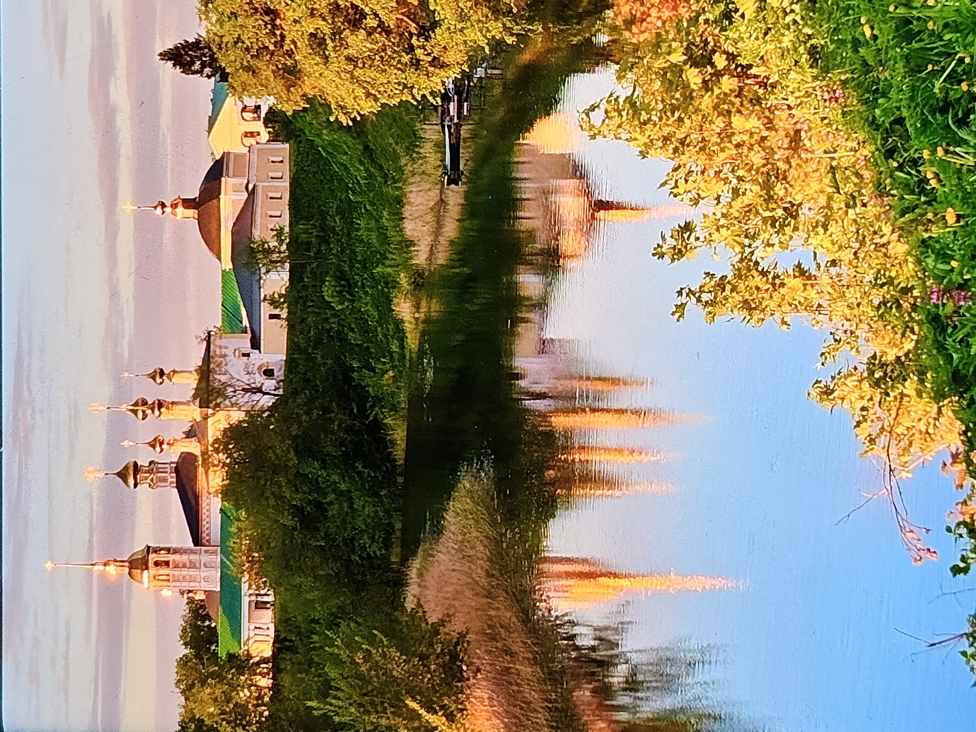 The image depicts a serene waterscape with a prominent religious complex in the background, likely situated in Vladimir, Russia. The architecture features multiple onion domes and steeples, characteristic of Russian Orthodox churches, bathed in the warm, golden light of sunset. The complex is situated on a gentle incline, surrounded by lush green trees and vegetation. A tranquil river or canal flows in the foreground, reflecting the illuminated structures and the soft hues of the sky. The water's surface is mostly calm, with a few ripples creating a distorted but beautiful mirror image of the scene above. In the water, near the right bank, two small boats are docked. The vegetation on the riverbank is dense, with reeds and grasses. The overall atmosphere is peaceful and picturesque, showcasing a blend of natural beauty and historical architecture.