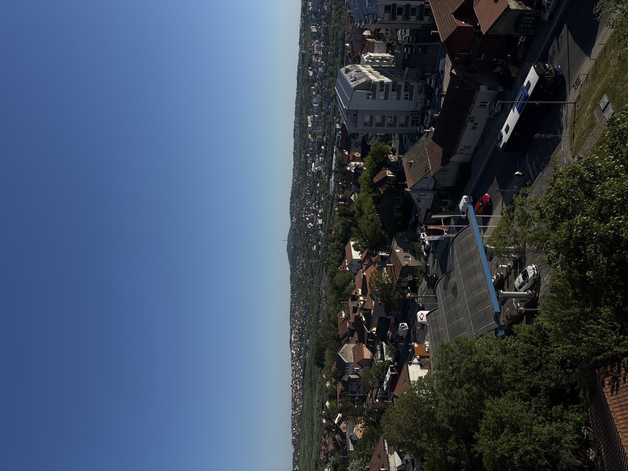 The image is an aerial view of a cityscape in Belgrade, Serbia, under a clear blue sky. Buildings, mostly residential, are densely packed and interspersed with green trees. A prominent feature is a curved, glass-roofed structure, possibly a gas station, surrounded by trees. Vehicles, including cars and a bus, are visible on the roads. In the distance, a tower or monument rises above the horizon. The scene is bathed in daylight, suggesting a sunny day.