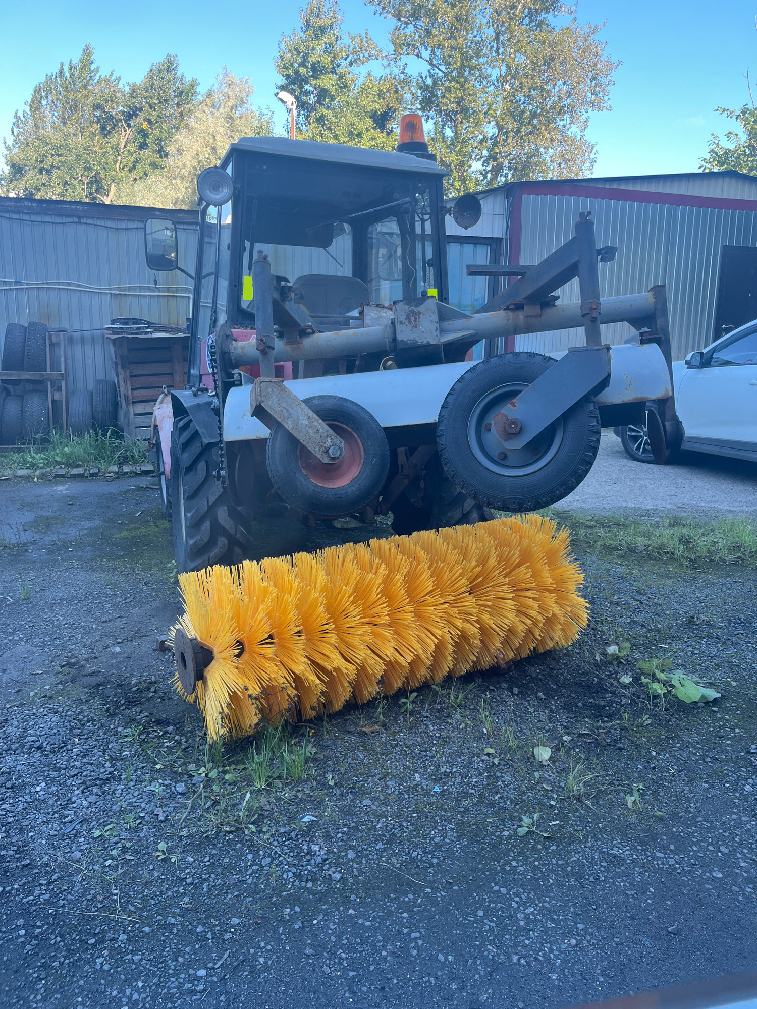 The image shows a street sweeping machine parked outdoors on a gravel surface. The machine, predominantly dark-colored, features a large, bright yellow brush attached to the front. The brush is angled downwards and appears to be the primary cleaning apparatus. Behind the brush, the vehicle has large, rugged tires and a metal frame. The cab of the machine is visible, with a warning light on top. In the background, there are industrial-looking buildings with corrugated metal siding, and trees. A white car is partially visible to the right. The scene suggests a service or maintenance area, possibly in an industrial or commercial part of Saint Petersburg, Russia, given the presence of the sweeping machine. The sky is clear and blue, indicating daytime.