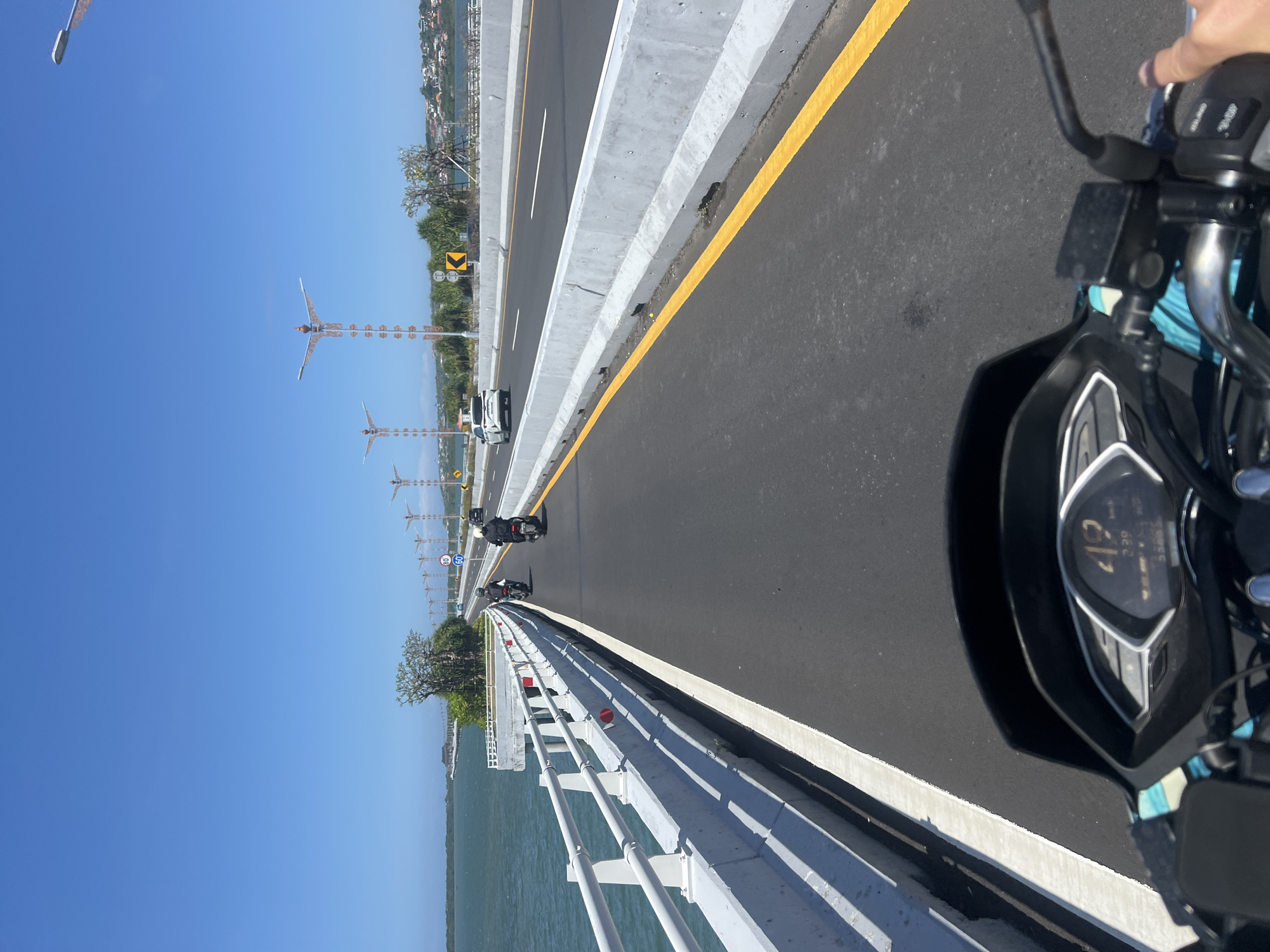 An over-the-shoulder shot captures a motorcycle's perspective as it travels on a bridge over water, likely in Kuta Selatan, Indonesia, given the tropical scenery and architectural style of the lamps lining the road. The rider's right hand grips the handlebar, and the motorcycle's digital dashboard displays a speed of 49 km/h. Two other motorcycles, each with a single rider, are ahead on the road. Further down the bridge, a white car is visible. The road is paved and has a yellow lane marker. On the left side of the road is a white railing, and beyond that, the turquoise water extends to a distant tree-lined shore. The sky is a clear, bright blue. The decorative lampposts, featuring intricate designs and bird-like figures, are characteristic of Balinese architecture, suggesting the bridge is a significant piece of infrastructure in the area, possibly connecting Nusa Dua to the mainland.
