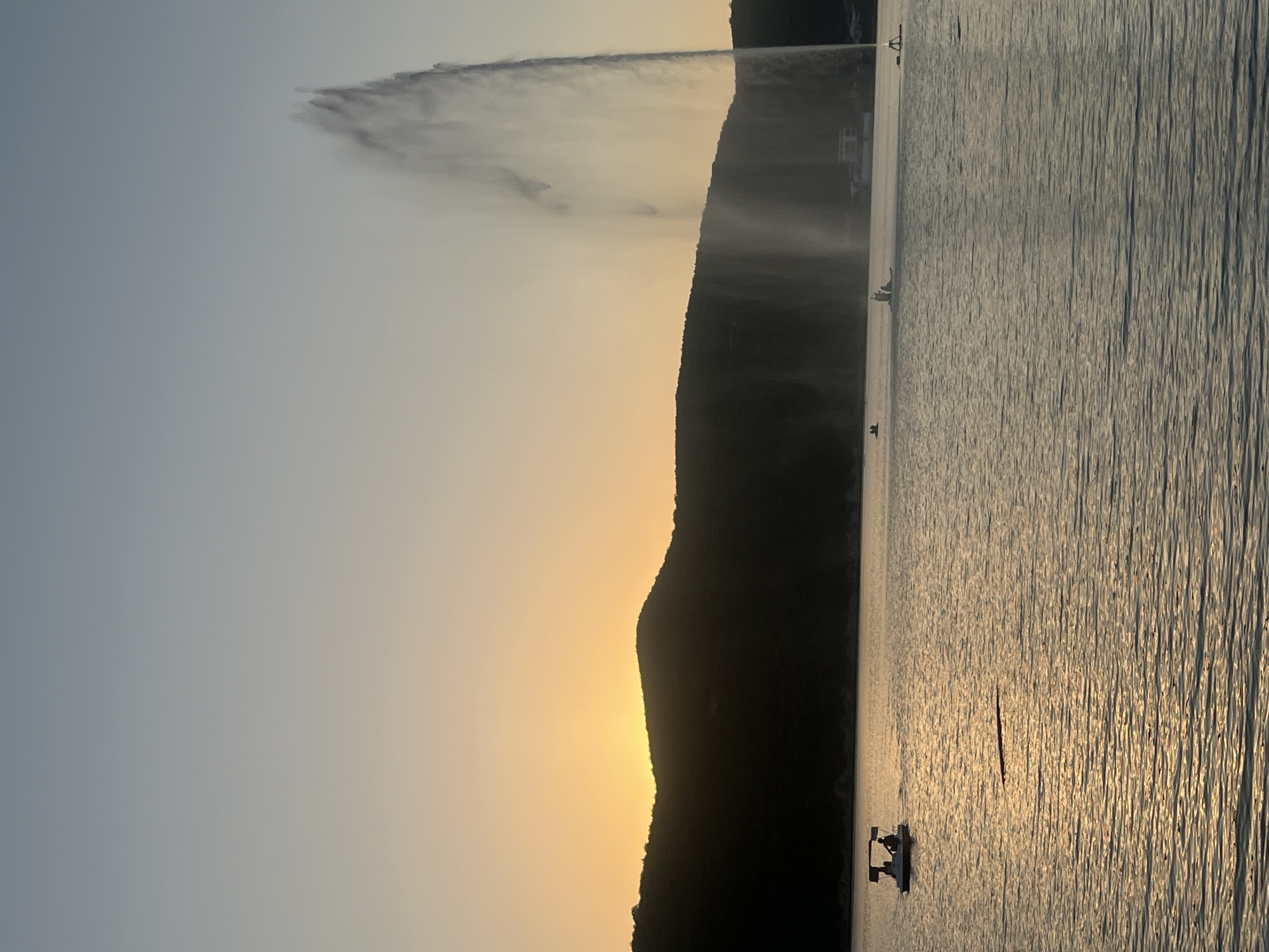 The image shows a large, wide fountain spraying water high into the air on a body of water. A dark, silhouetted mountain range forms the horizon behind the water. The sky is a gradient of pale blue at the top fading into a warm yellow-orange near the horizon, indicating either sunrise or sunset. On the water, a small pedal boat with at least two people is visible in the left foreground, and several other small boats are scattered further out on the water. The water itself is rippled and reflects the warm light from the sky. The overall scene depicts a serene, recreational setting on a lake or large reservoir. This fountain is a prominent landmark at the Abrau-Durso wine estate in Abrau-Dyurso, Russia, and is known for its impressive height and scale.