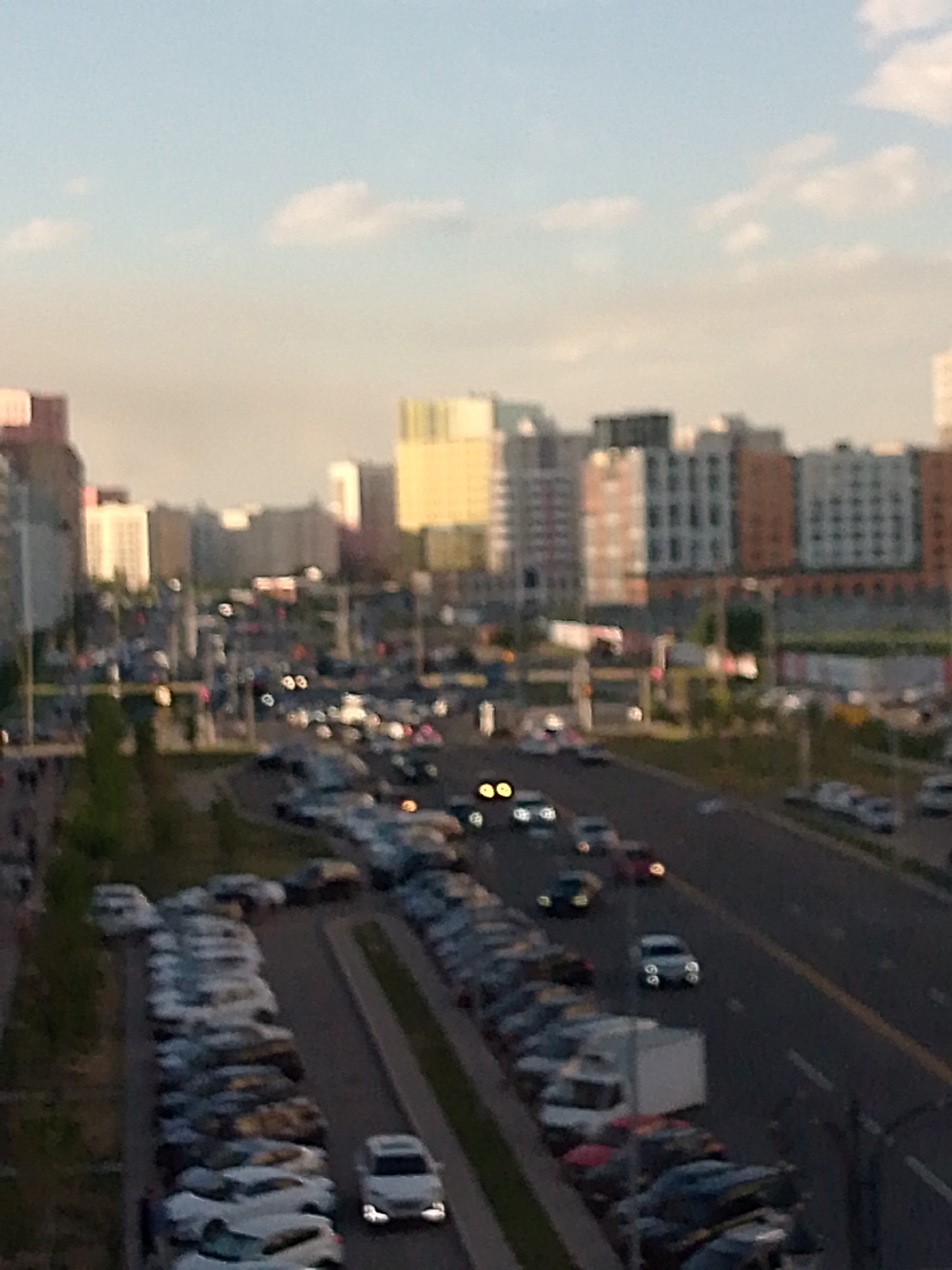 This is a blurry, horizontal, medium-distance shot of an urban scene, likely taken from a high vantage point, such as a building or bridge. The time of day appears to be late afternoon or early evening, as suggested by the soft, diffused sunlight and the presence of car headlights. The sky is a pale blue with a few wispy clouds, and the overall lighting is warm.
The main subjects are a busy street and a large parking lot filled with numerous cars. The street is lined with parked vehicles and appears to have traffic moving in both directions, indicated by the glowing headlights and taillights of some cars. A few vehicles are clearly visible, including a white van or truck in the parking lot and a light-colored car with its headlights on, driving on the street. The parking lot is densely packed with a variety of cars, mostly dark in color, arranged in rows.
In the background, a collection of multi-story buildings forms the cityscape. These buildings are varied in architectural style and color, with some appearing to be residential or office blocks. The colors range from light beige and white to darker bricks and gray facades. One building has a distinctive yellow facade.
The scene depicts a typical urban environment with elements of daily life, such as commuting and parking. There is no indication of any specific event or emotional tone, beyond the general activity of a city. No visible text is present in the image.