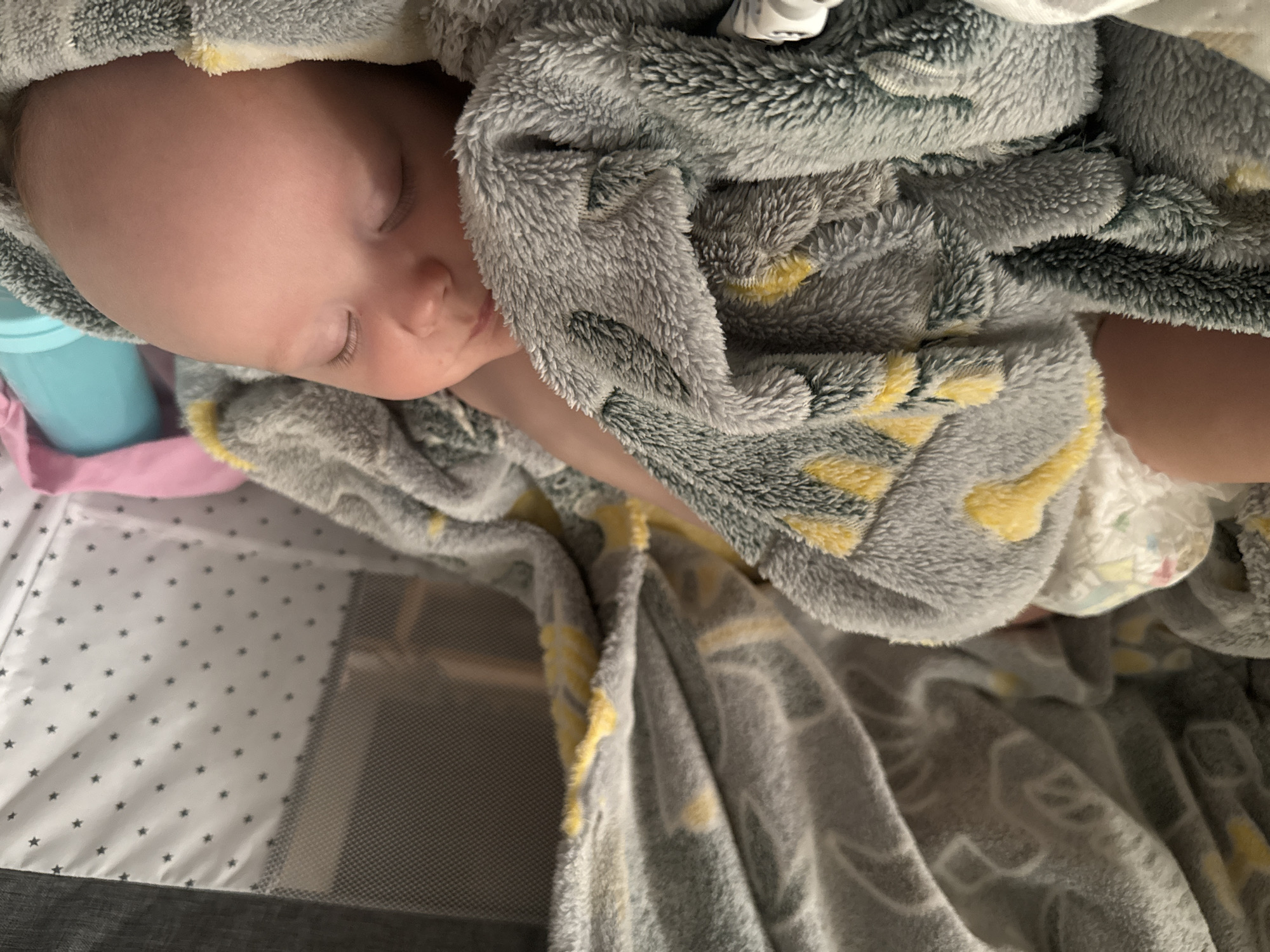 This image depicts an indoor scene, understood to be located in the city of Moscow, Russia. A light-skinned infant is observed sleeping peacefully on their right side within a portable playpen or travel crib. The infant's eyes are closed, and their head, with sparse light hair, rests on the soft surface. A plush, grey blanket with yellow and white geometric or abstract patterns partially covers the infant's body, revealing a portion of a bare leg or arm and a white patterned diaper. The interior of the playpen features a white fabric lining adorned with small black star patterns. A mesh side panel is visible to the left. In the background, near the infant's head, a blue lidded bottle and a piece of pink fabric are visible against the starry lining. The lighting is soft, creating a calm and intimate atmosphere.