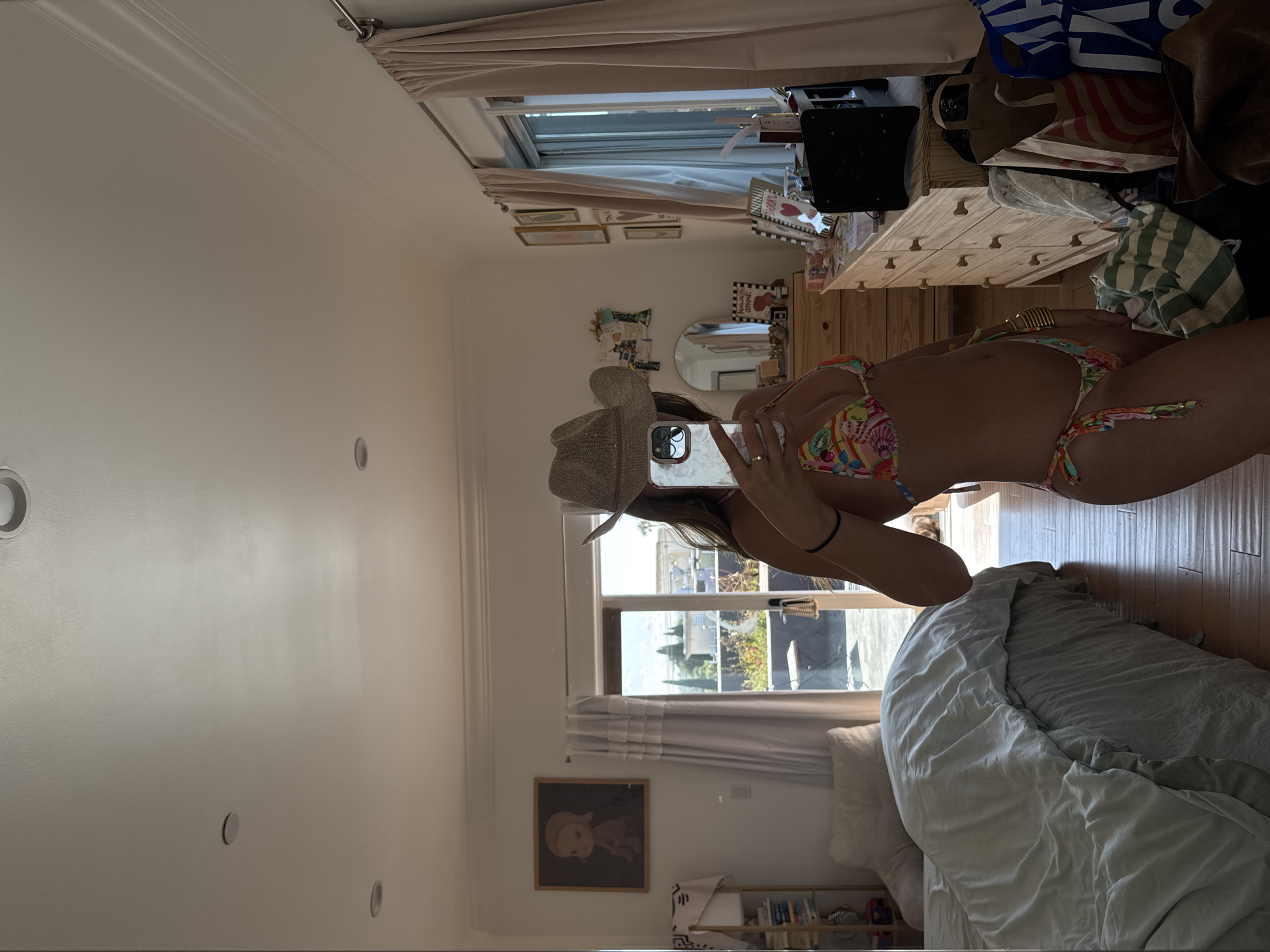 A mirror image shows a woman in a bikini and a cowboy hat holding a cell phone in front of her face, taking a photo. She is standing in a bedroom. In the background, through a window, is a view typical of Los Angeles: palm trees and other greenery are visible. The scene includes furniture, a bed, and decor items within the bedroom setting.