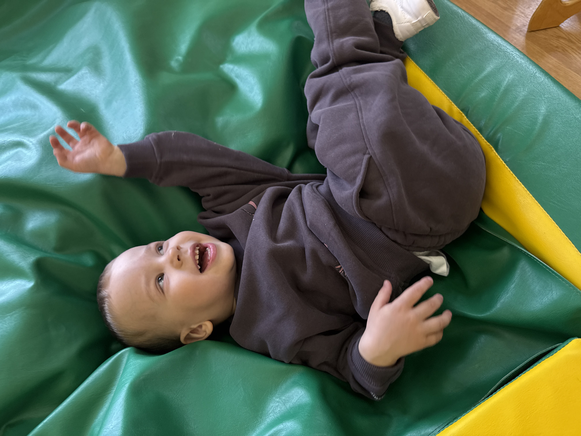 A child, wearing a dark-colored sweatshirt and pants, lies on a green and yellow mat. The child is on their back with their legs raised in the air and their hands outstretched. The child's mouth is open in a smile or laugh. The location is not possible to be determined based on visual information.