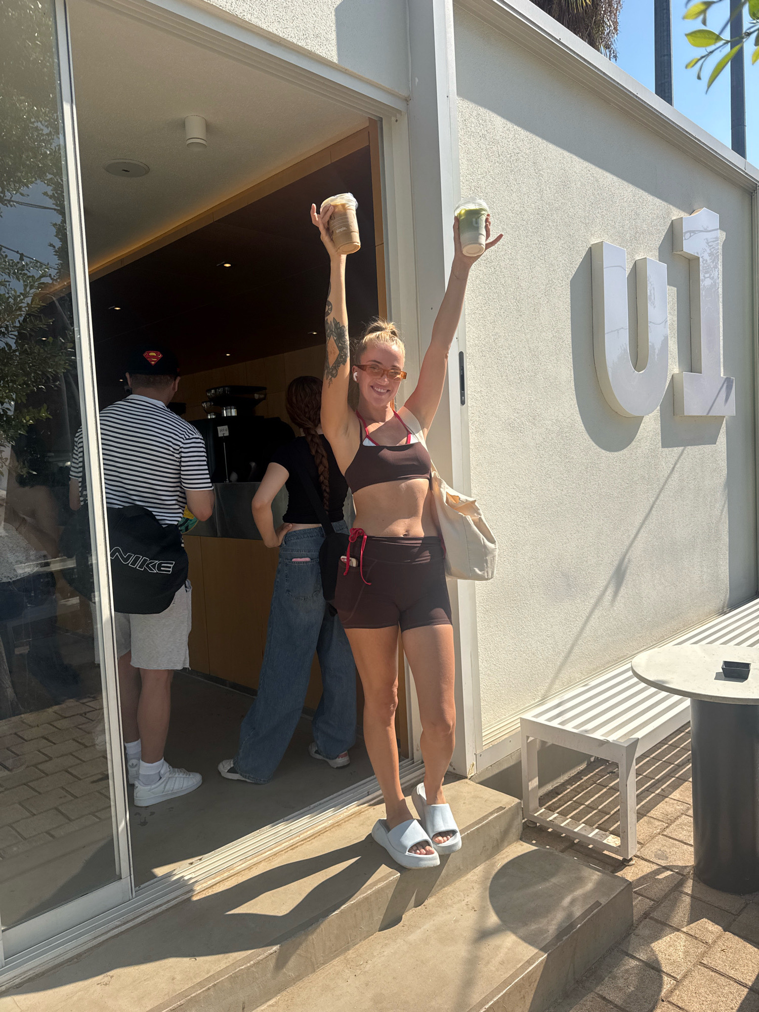 A young adult female stands on the steps outside a building with a large "U1" logo, holding up two drinks. She wears a brown sports bra, brown shorts, and light blue slides. She has blonde hair pulled back and is wearing sunglasses. Behind her, a man in a striped shirt and shorts and another person in a black shirt and jeans are inside the building. A white tote bag is slung over the woman's shoulder. The scene is outdoors on a sunny day, with shadows cast on the concrete ground and a table and bench visible to the right. The architectural style and the "U1" logo suggest a modern establishment, possibly a cafe or shop. The presence of palm trees in the background and the bright sunlight are consistent with a Mediterranean climate like that found in Antalya, Turkey.