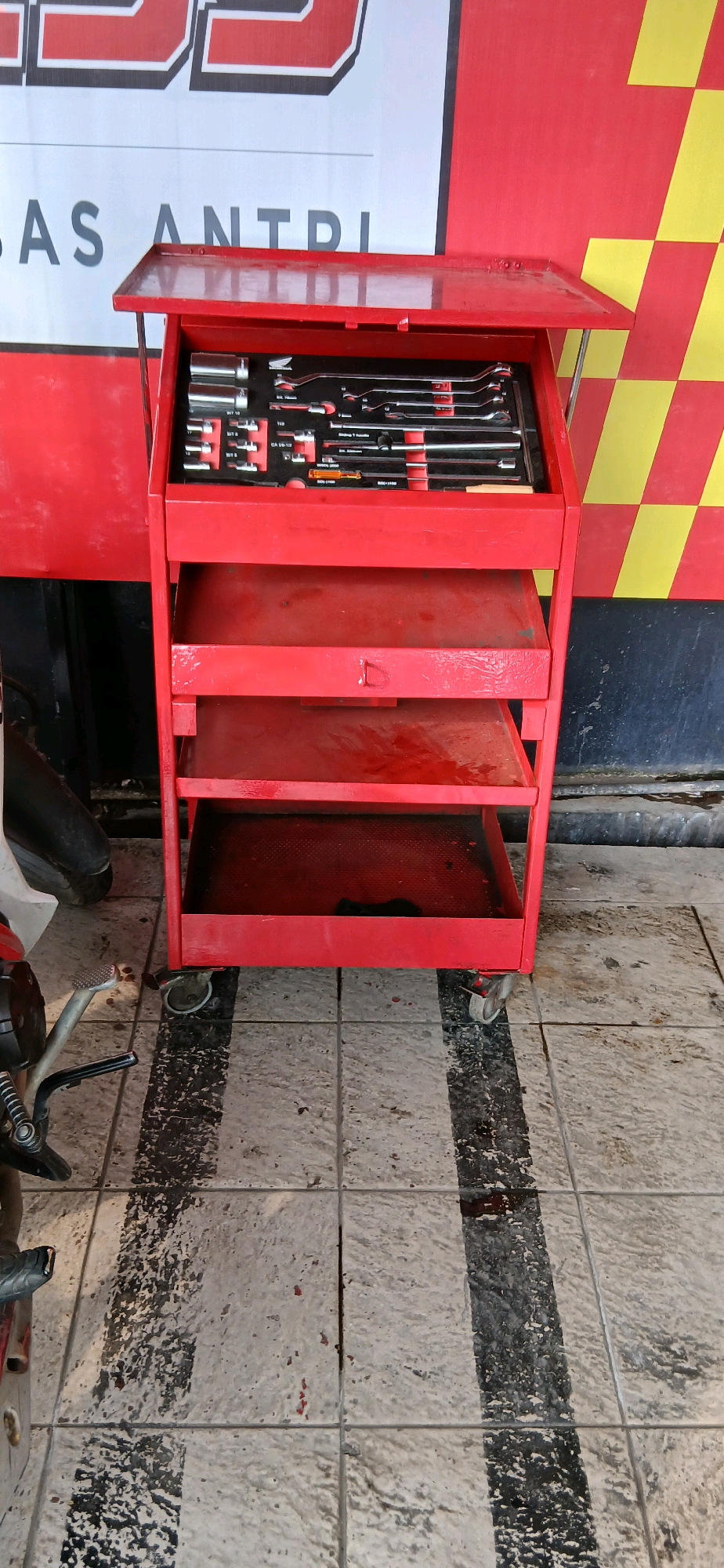 An open, red, rolling tool cart is positioned on a tiled floor. The top section of the cart, which is angled upwards, contains an assortment of wrenches, sockets, and other hand tools neatly organized in black foam cutouts. Below this top section are two empty shelves. The tool cart is placed in front of a wall that is red on the left and features a red and yellow checkered pattern on the right. A portion of a motorcycle, including its front wheel and part of its frame, is visible to the left of the tool cart. The floor is tiled in a light color with alternating wide black stripes that appear worn and marked with dark stains. Red paint splatters are also visible on the floor tiles. This scene is likely located in a workshop or garage setting, possibly in Samarinda, Indonesia, given the context provided. There are no people visible in the image.