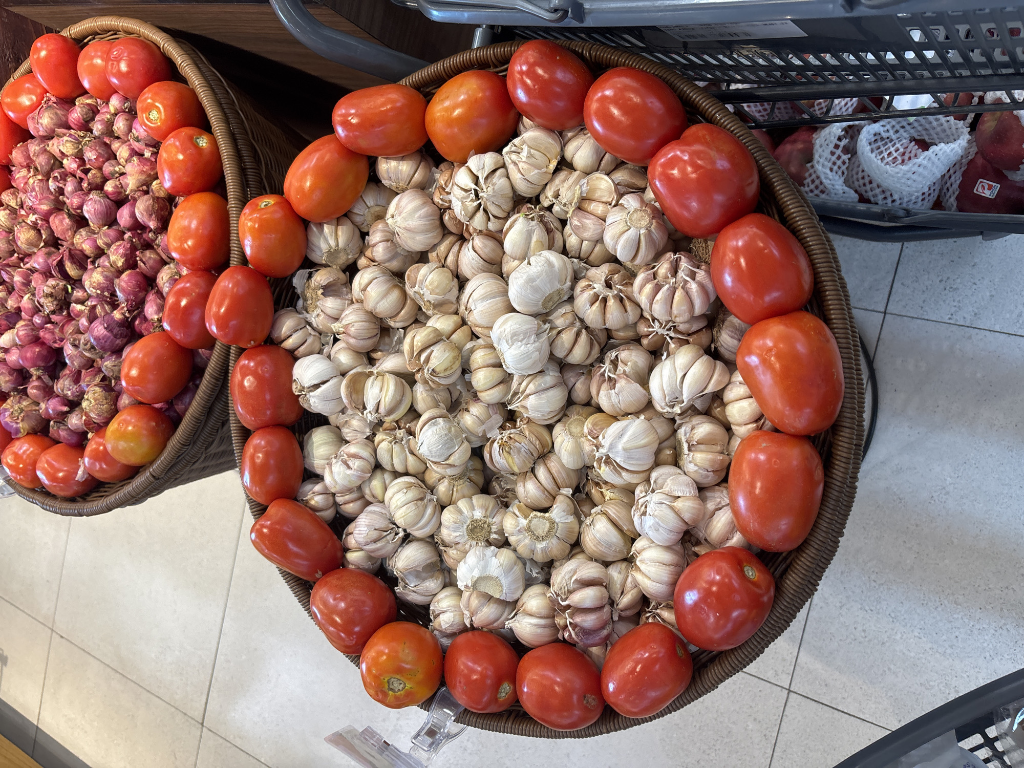 Two woven baskets are displayed on a tiled floor. The upper basket contains numerous small, red onions and some red tomatoes. The lower basket is filled with heads of garlic and red tomatoes. The tomatoes in the lower basket are larger and more prominent, some arranged around the perimeter of the basket, while the garlic is clustered in the center. The lighting suggests an indoor environment, possibly a market or grocery store. The location is Kuta Selatan, Indonesia.