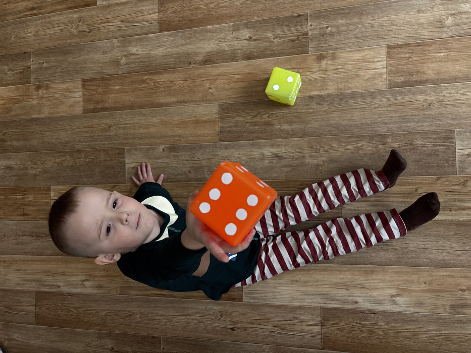 An overhead photograph captures a young male child seated on a wooden floor in an indoor residential setting in the city of Pavlovsky, Russia. The child, with light skin and short light brown hair, is dressed in a dark green long-sleeved shirt with a cream-colored collar and horizontal red and white striped pants, along with dark brown socks. He faces forward, looking directly at the camera with a neutral expression. His right arm is extended towards the camera, holding a large, bright orange six-sided die, which prominently displays faces with five and two white pips. A second large, neon green six-sided die, showing faces with four and two white pips, rests on the light brown wooden plank floor to the child's right.