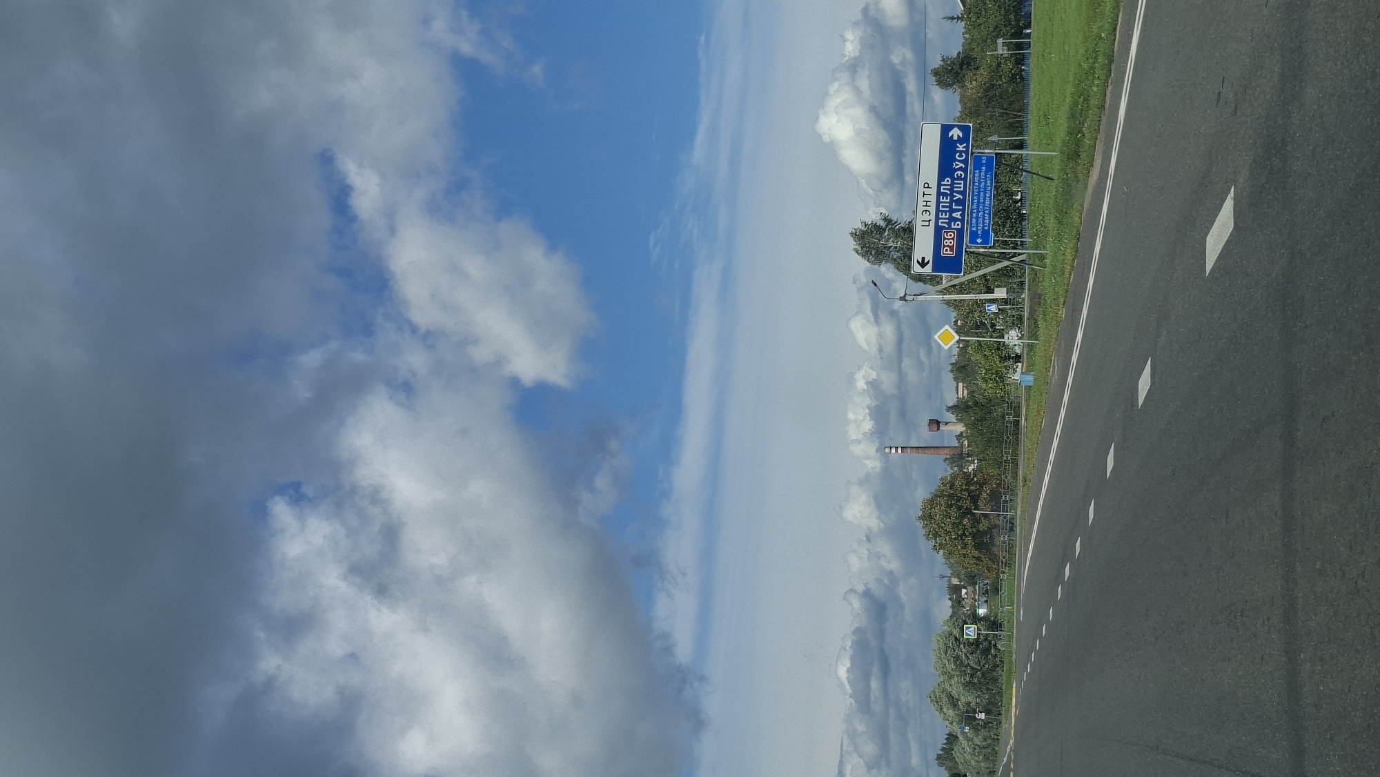 The image depicts a road scene in the city of Myadzyel, Belarus. A two-lane asphalt road with white dashed markings runs diagonally from the bottom left to the top right of the frame. On the right side of the road, a blue directional sign indicates "Цэнтр" (Center) with an arrow pointing left, and "ЛЕПЕЛЬ БАГУШЭУСК" (Lepel Bagusheusk) with an arrow pointing right. Below this, another blue sign shows "ДЗРЖАУНАЯ УСТАНОВА" (State Institution) with an arrow pointing left and "МІНСК-ГАЙНА-ЛАГОДА-65" (Minsk-Gaina-Logoda-65) with an arrow pointing right, and "АДЗАРУЛЮЦЫЯ ЦЭНТР" (Adarulyutsya Center) with a left arrow. A yellow diamond-shaped sign and a white square sign with a blue border are also visible on the right. The landscape features a line of trees and vegetation along the roadside, with industrial structures, including a tall brick chimney, visible in the background. The sky is partly cloudy with a mix of dark and light clouds against a blue sky.