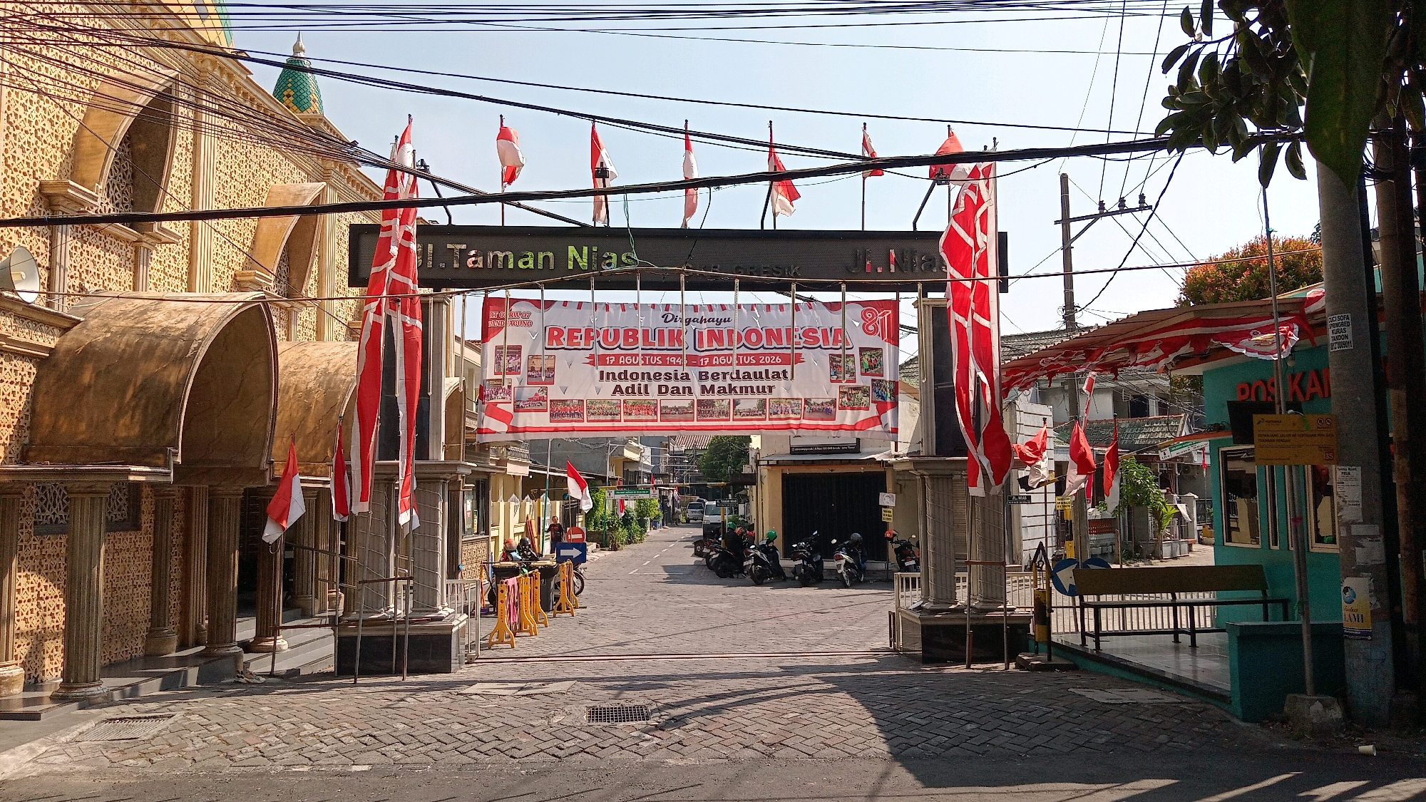 The image depicts a street scene in Randuagung, Indonesia, adorned with red and white Indonesian flags and banners celebrating Republic Day. A large banner reads "DIRGAHAYU REPUBLIK INDONESIA 81" with dates "17 AGU TUS 1945 - 17 AGU TUS 2026" and the slogan "Indonesia Berdaulat Adil Dan Makmur". Several smaller Indonesian flags are hung across the street and attached to buildings.
On the left, a building with intricate patterned walls and arched entrances is visible. Flags hang prominently in front of this building. To the right, a turquoise building with the text "POS KAI" (Indonesian Post Office) is seen, also decorated with flags and banners.
The street itself is paved with cobblestones and is lined with several motorcycles, some parked and others being ridden by people wearing helmets. A few individuals are visible walking or standing near the buildings. In the background, more buildings and trees can be seen under a clear sky. Electrical wires crisscross the upper portion of the image. The scene appears to be a public space during a festive occasion, likely a commemoration of Indonesian Independence Day.