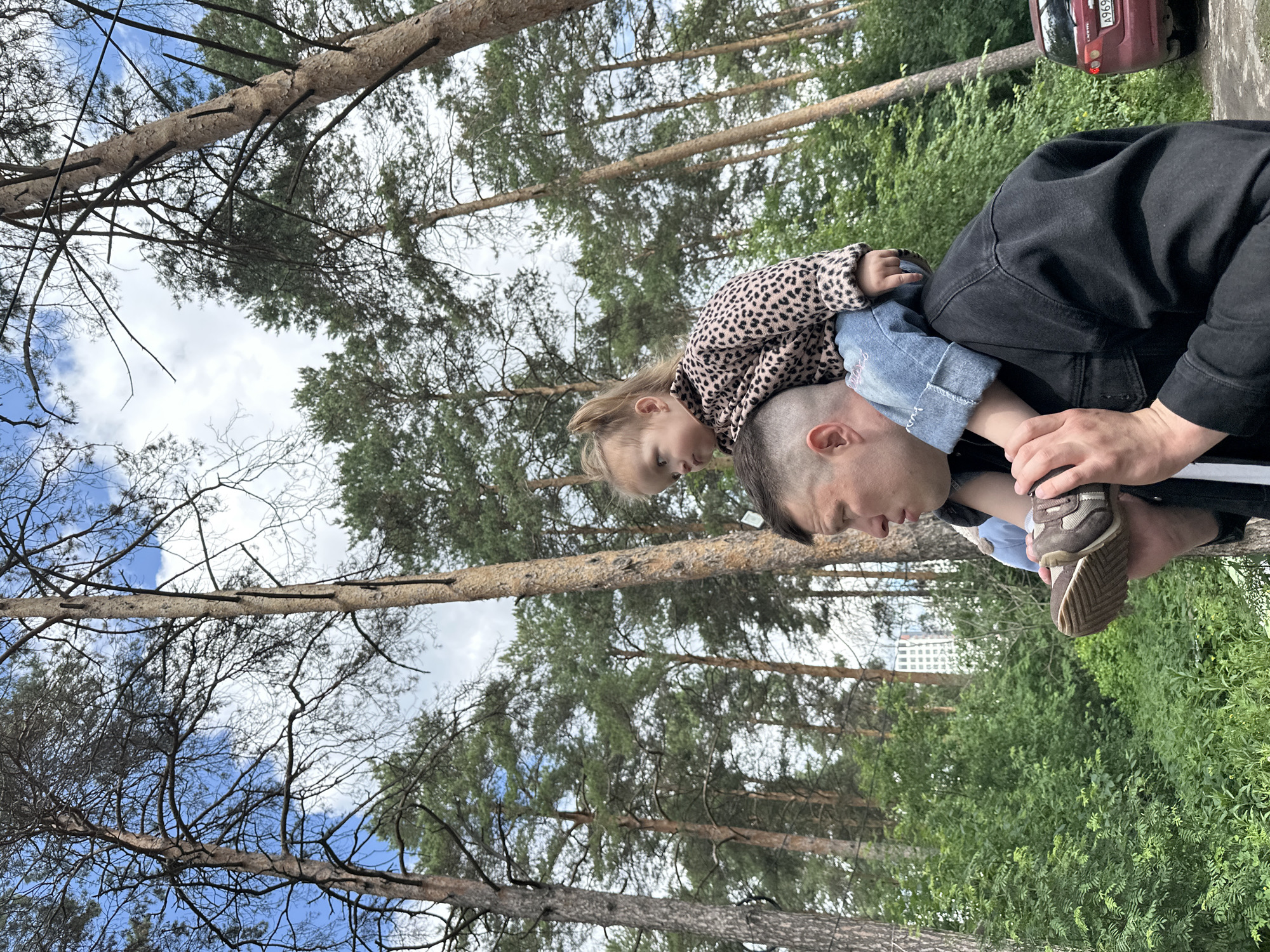 A father carries his young daughter on his shoulders in a forest. The daughter, facing left, wears a leopard-print jacket and blue jeans. The father, wearing a black jacket, looks to the left. In the background, tall pine trees dominate the scene, with a blue sky and white clouds above. A red car is partially visible on the right side of the image, and a distant building can be seen through the trees. The image appears to have been taken in Yekaterinburg, Russia, likely in a park or forested area on the outskirts of the city.