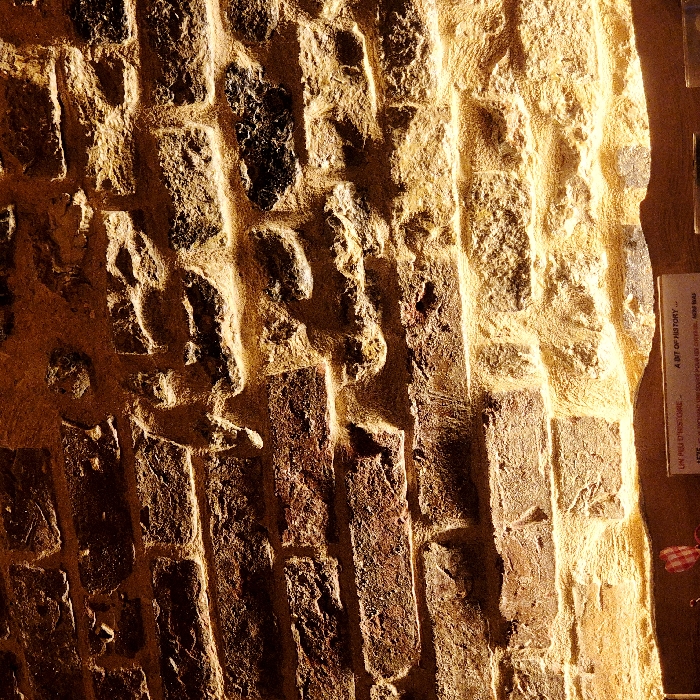 A close-up depicts a weathered brick wall, bathed in warm, golden light. The bricks vary in color, from dark brown to reddish hues. Thick, light-colored mortar fills the gaps between the bricks. On the right, a sign with faded text is partially visible. The rough texture of the wall suggests age and history.