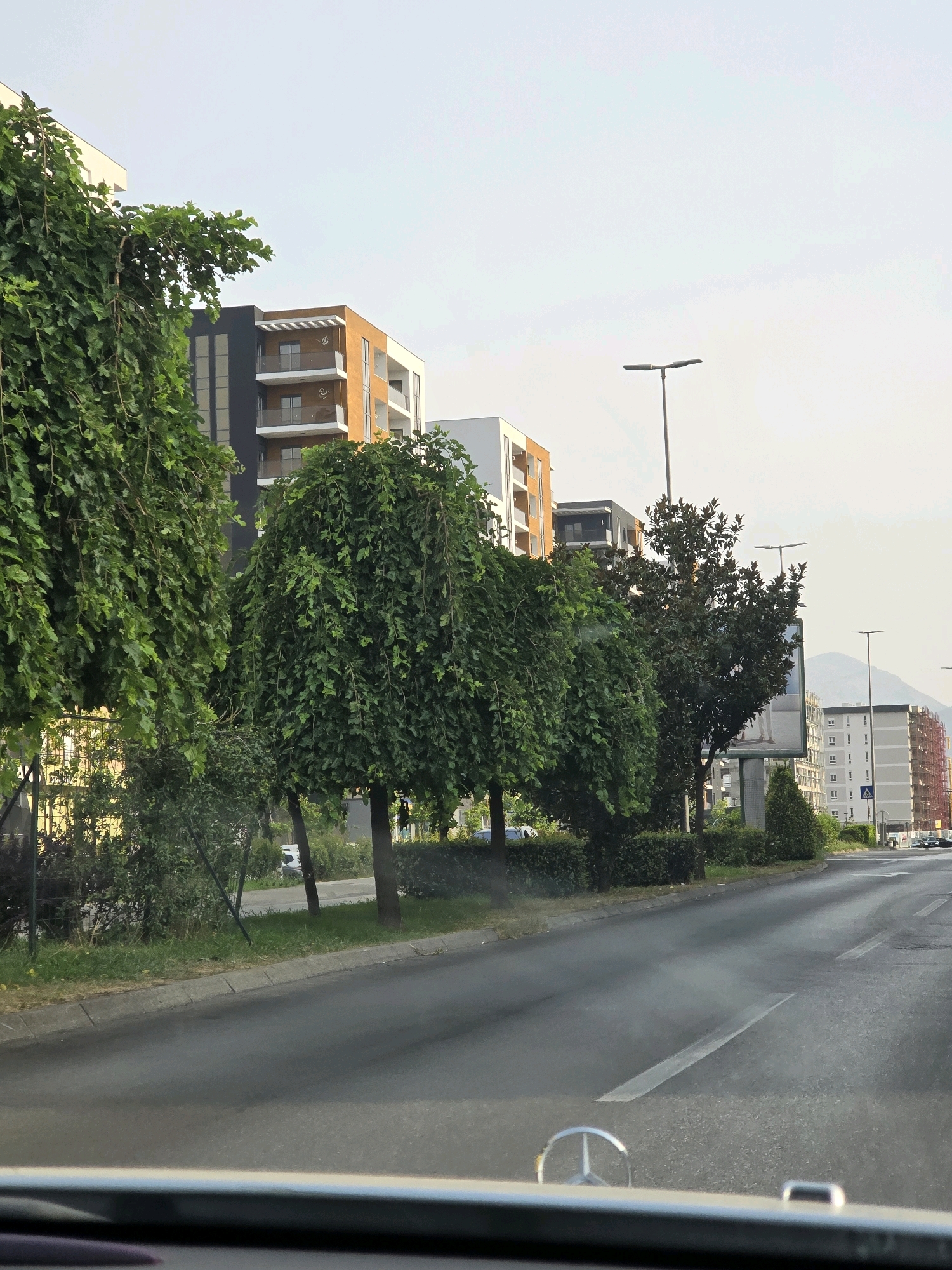 The image depicts a street scene in Podgorica, Montenegro, on a clear day with a light haze. The perspective is from inside a vehicle, looking forward. The foreground shows the dashboard of a car, including a silver Mercedes-Benz emblem. A road with painted lane markings stretches into the distance. Lined along the right side of the road are several modern apartment buildings, characterized by contemporary architecture with balconies and varied facades, some featuring orange and white elements, others white and dark grey. To the left of the road, several large, lush green trees with drooping branches are prominent, obscuring some of the buildings behind them. Further down the street, more apartment buildings and some construction can be seen. Streetlights are visible at intervals along the road. In the background, mountains are faintly visible against the pale sky. The overall impression is an urban environment with a mix of residential architecture and abundant greenery.