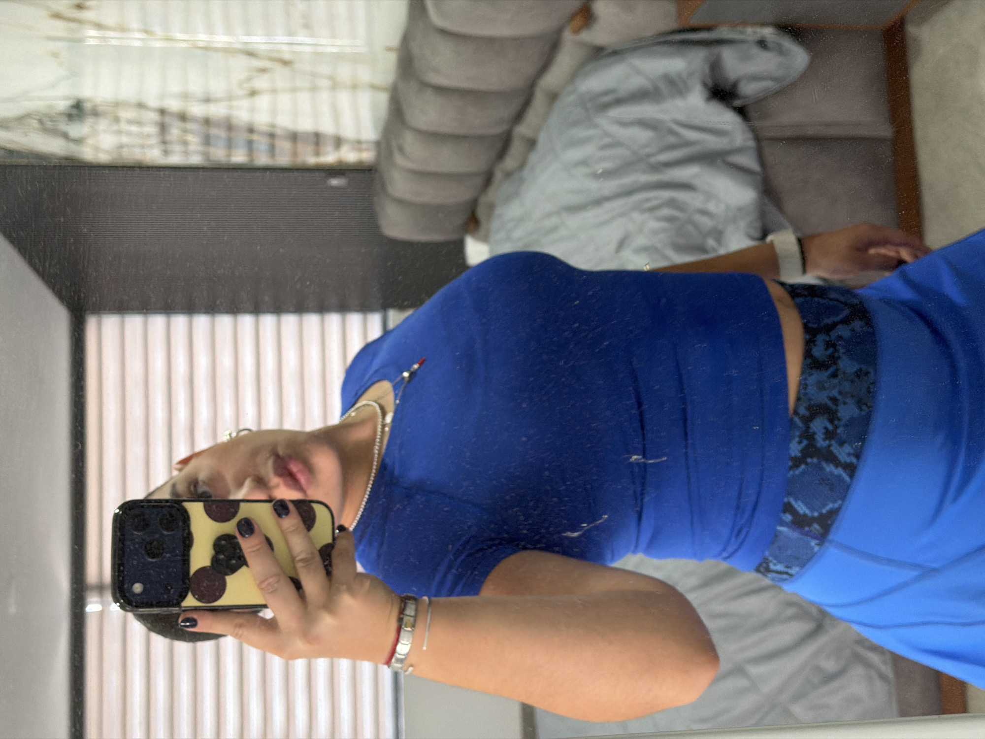 A person is reflected in a mirror, holding a phone in their left hand to take a picture. They are wearing a blue top, blue bottoms, and a watch on their right wrist. A necklace is visible around their neck. Behind them is a padded headboard and a window with blinds. The location is assumed to be indoors, possibly a residential setting, in the city of Puebla, Mexico.